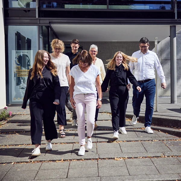 Eine Gruppe von sieben Personen, die in lässiger Kleidung und weißen Sneakers, fröhlich eine Treppe hinuntergeht. Im Hintergrund ist ein modernes Gebäude sichtbar.