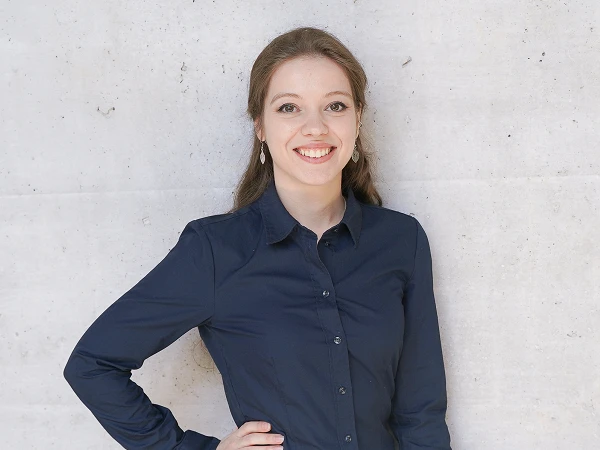 Eine lächelnde junge Frau steht mit einer Hand in die Hüfte gestützt vor einer grauen Wand. Sie trägt ein dunkles Hemd und hat ihre Haare offen.