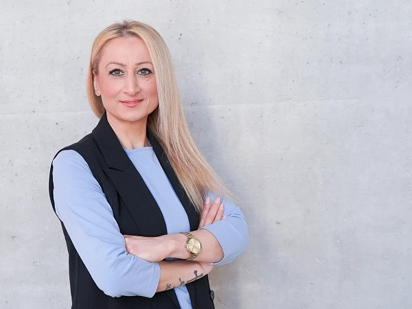 Eine Person mit langen blonden Haaren steht mit überkreuzten Armen vor einer grauen, strukturierten Wand. Sie trägt ein hellblaues Oberteil und einen schwarzen Blazer, wobei sie freundlich in die Kamera lächelt.