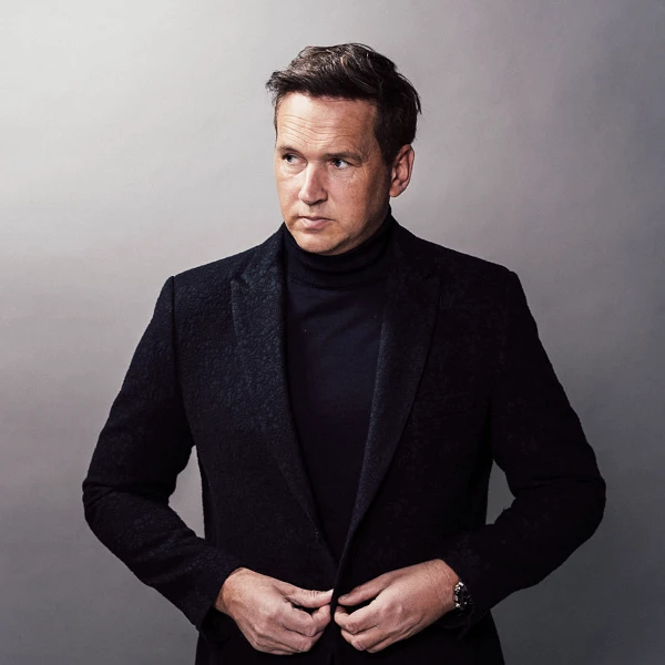 Ein Mann in einem eleganten schwarzen Anzug mit einem Rollkragenpullover steht vor einem neutralen Hintergrund und schaut nachdenklich in die Ferne, während er seinen Anzug zurechtzupft.