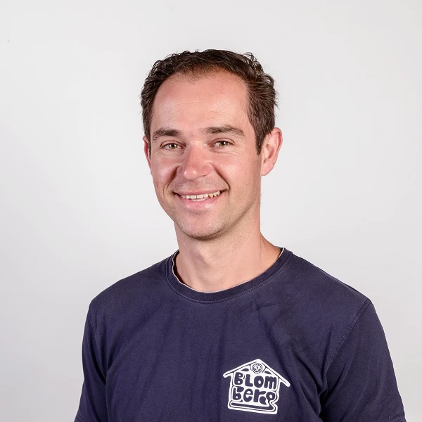 Ein lächelnder Mann mit kurzen, dunklen Haaren trägt ein dunkelblaues T-Shirt mit einem Logo auf der linken Brustseite und steht vor einem einfarbigen Hintergrund.