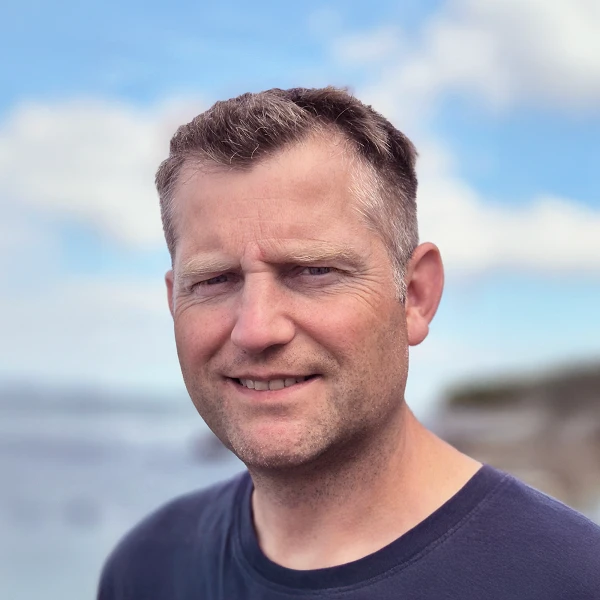 Ein Mann mit kurzen Haaren und einem freundlichen Lächeln steht vor einem verschwommenen Hintergrund von Wasser und Himmel.