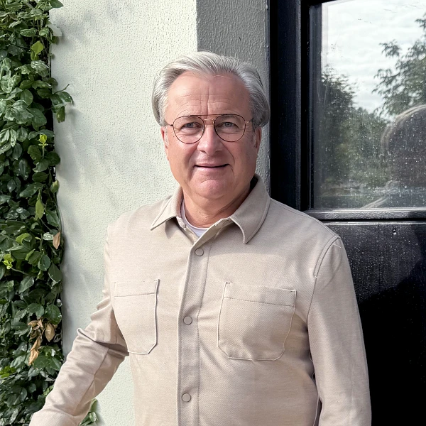 Ein lächelnder Mann mit grauen Haaren und Brille steht an einer Wand, die mit grünen Pflanzen bewachsen ist. Er trägt ein beiges Hemd mit zwei Brusttaschen. Im Hintergrund sieht man einen Fensterrahmen und einen leicht bewölkten Himmel.