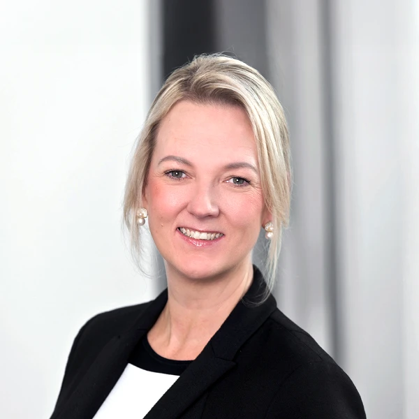 Eine lächelnde Frau mit blonden Haaren trägt einen schwarzen Blazer und eine weiße Bluse, vor einem hellen, unscharfen Hintergrund.