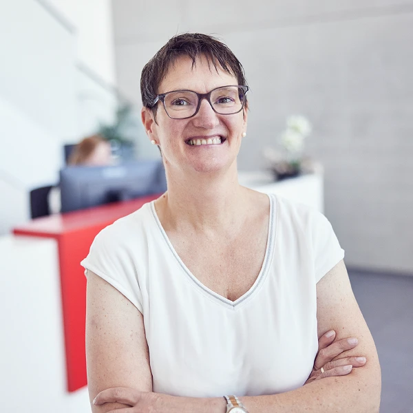 Eine lächelnde Frau mit kurzen, braunen Haaren und Brille steht mit verschränkten Armen in einem modernen Empfangsbereich, während sie in die Kamera schaut. Im Hintergrund ist ein Empfangstresen sichtbar.