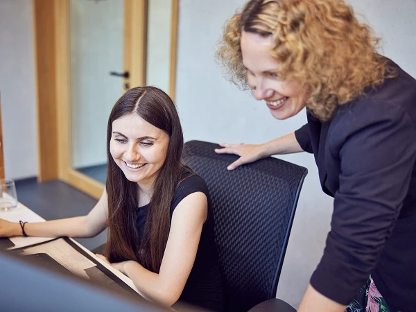 Zwei Frauen sitzen an einem Schreibtisch, eine von ihnen zeigt auf den Computerbildschirm, während die andere lächelt und aufmerksam zusieht. Im Hintergrund ist ein Büro mit Holz- und Glaselementen erkennbar.