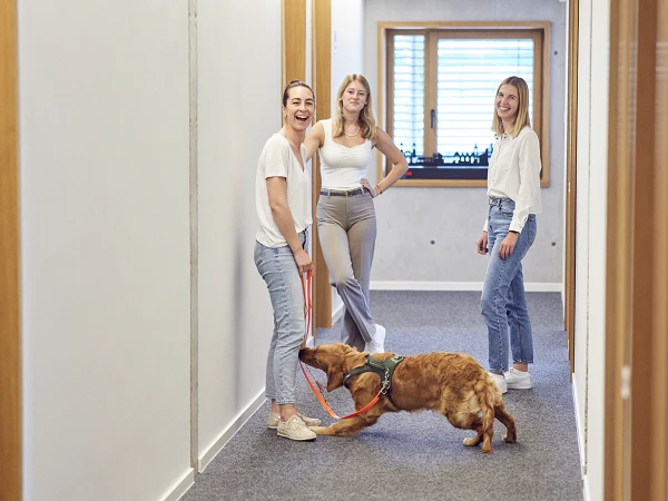 Drei junge Frauen stehen in einem hellen Flur, während ein Hund an einer Leine zieht und auf den Boden schaut. Die Frauen lachen und scheinen eine angenehme Zeit miteinander zu verbringen.