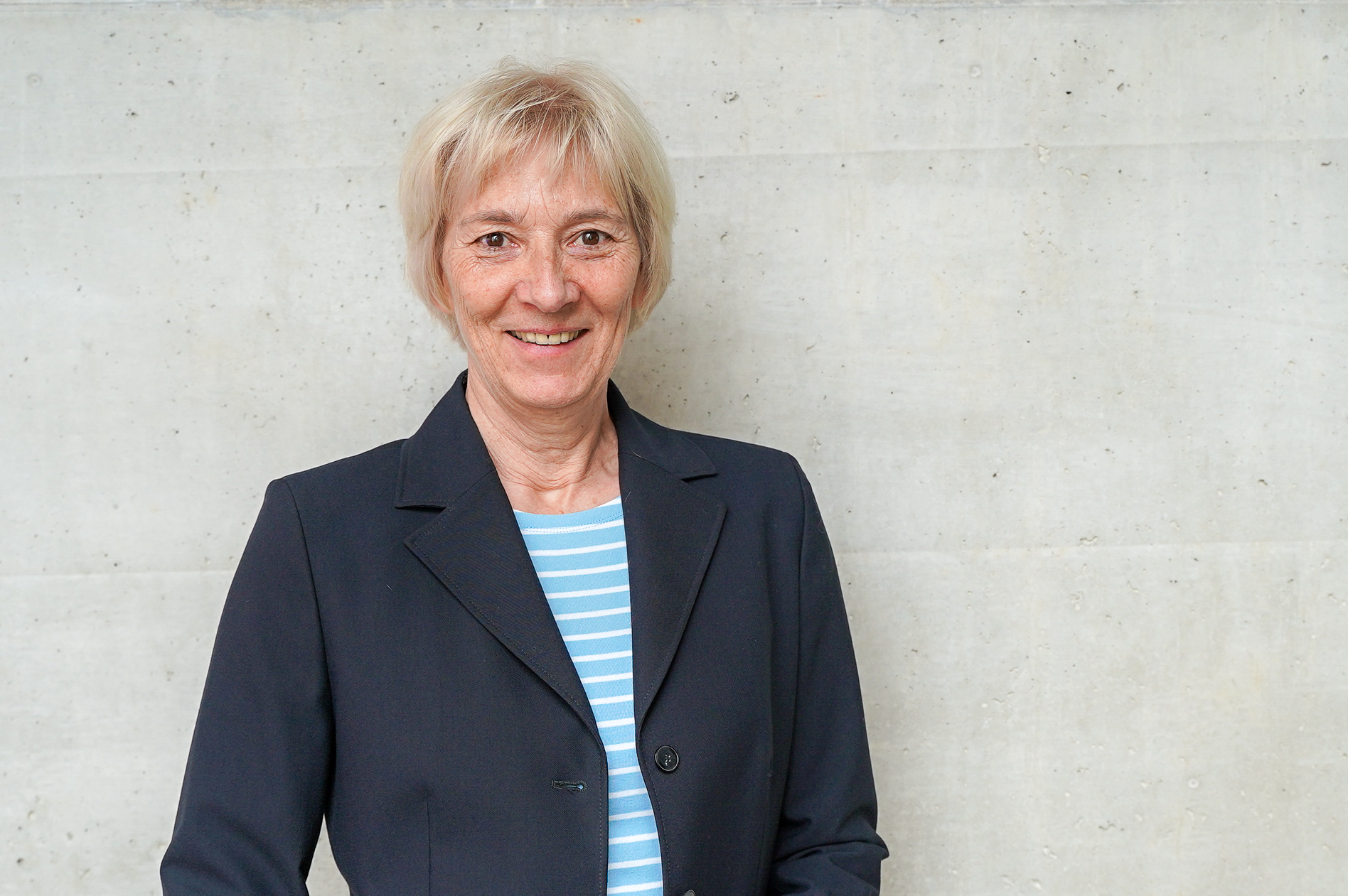 Die Frau mit blonden, kurzen Haaren trägt einen schwarzen Blazer über einem gestreiften Shirt und lächelt vor einer neutralen, grauen Wand.