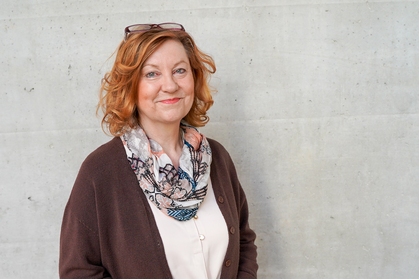 Eine lächelnde Frau mit lockigem, rotem Haar und einer bunten Schal steht vor einer grauen Wand. Sie trägt einen braunen Cardigan und hat eine Brille auf dem Kopf.