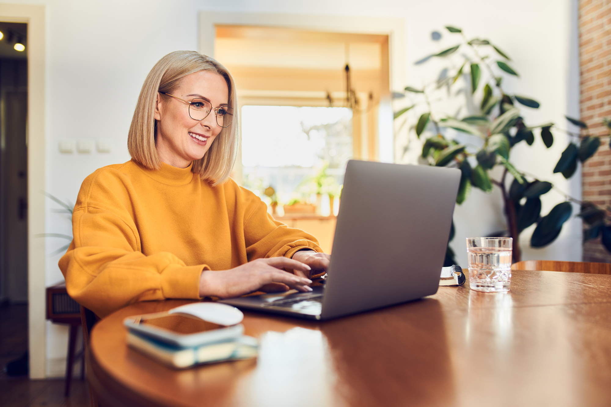 Eine lächelnde Frau mit blonden, schulterlangen Haaren sitzt an einem Holztisch und arbeitet am Laptop. Sie trägt einen orangefarbenen Pullover und eine Brille. Im Hintergrund sind Pflanzen und ein helles Raumambiente zu sehen. Ein Wasserglas und ein Notizbuch liegen auf dem Tisch.