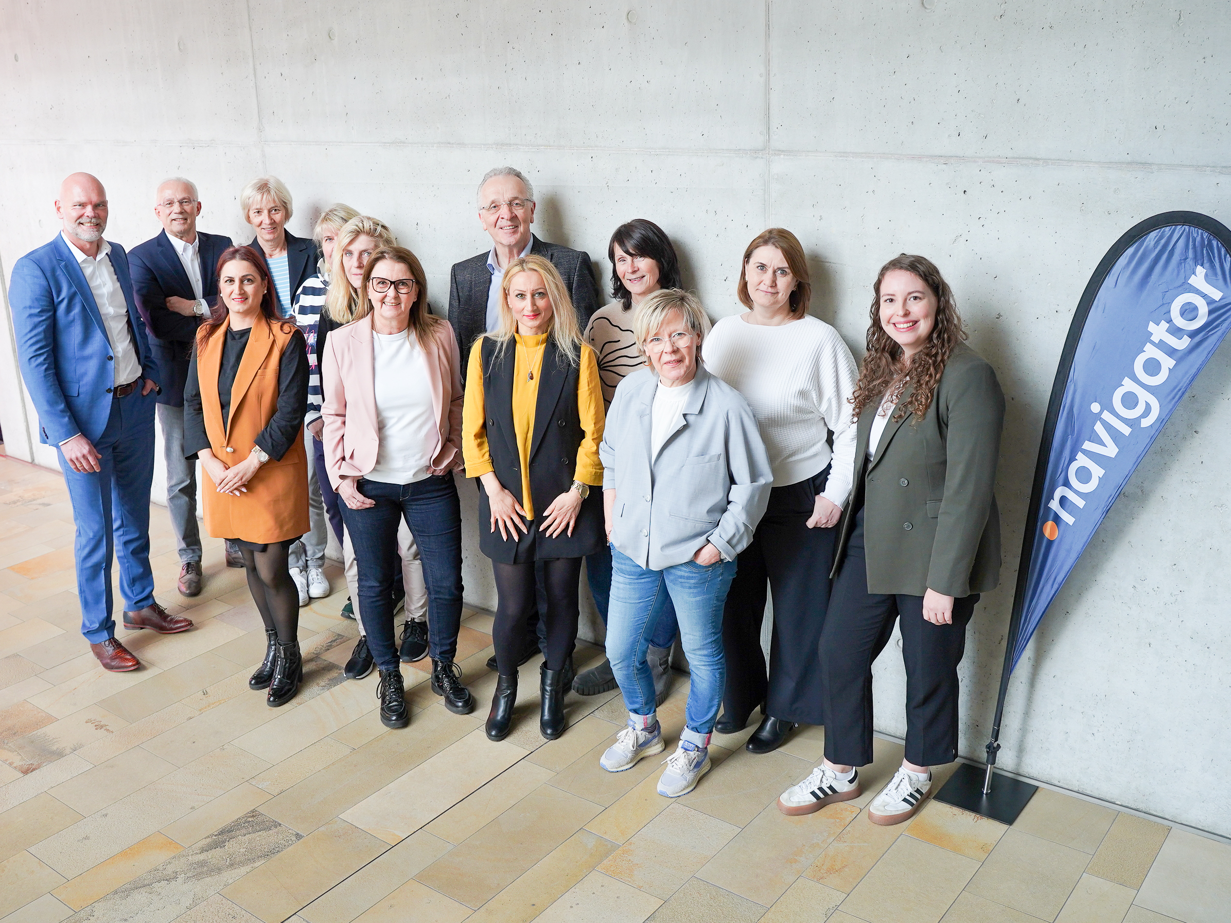 Eine Gruppe von 14 Personen steht vor einer grauen Wand, einige in formeller Kleidung, andere leger gekleidet. Rechts im Bild befindet sich ein Banner mit der Aufschrift „navigator“. Die Gruppe lächelt freundlich in die Kamera.