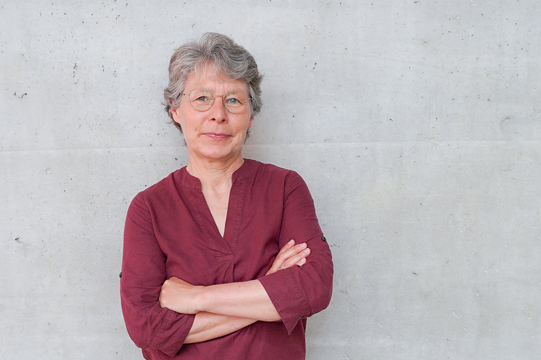 Eine Frau mit kurzen grauen Haaren und Brille steht mit verschränkten Armen vor einer grauen Betonwand, sie trägt ein rotes Oberteil und schaut direkt in die Kamera.