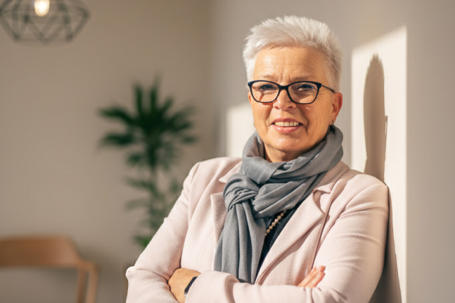 Eine lächelnde ältere Frau mit kurzen, grauen Haaren und einer Brille steht lässig mit verschränkten Armen da. Sie trägt einen hellen Blazer und einen grauen Schal, und im Hintergrund ist eine Pflanze sowie ein modernes Lichtobjekt sichtbar.