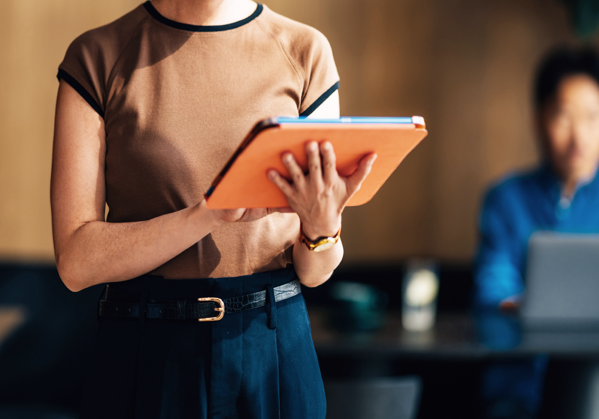 Eine Person hält ein orangefarbenes Tablet in den Händen, während sie in einem modernen Café steht; im Hintergrund ist eine weitere Person zu sehen, die an einem Laptop arbeitet.