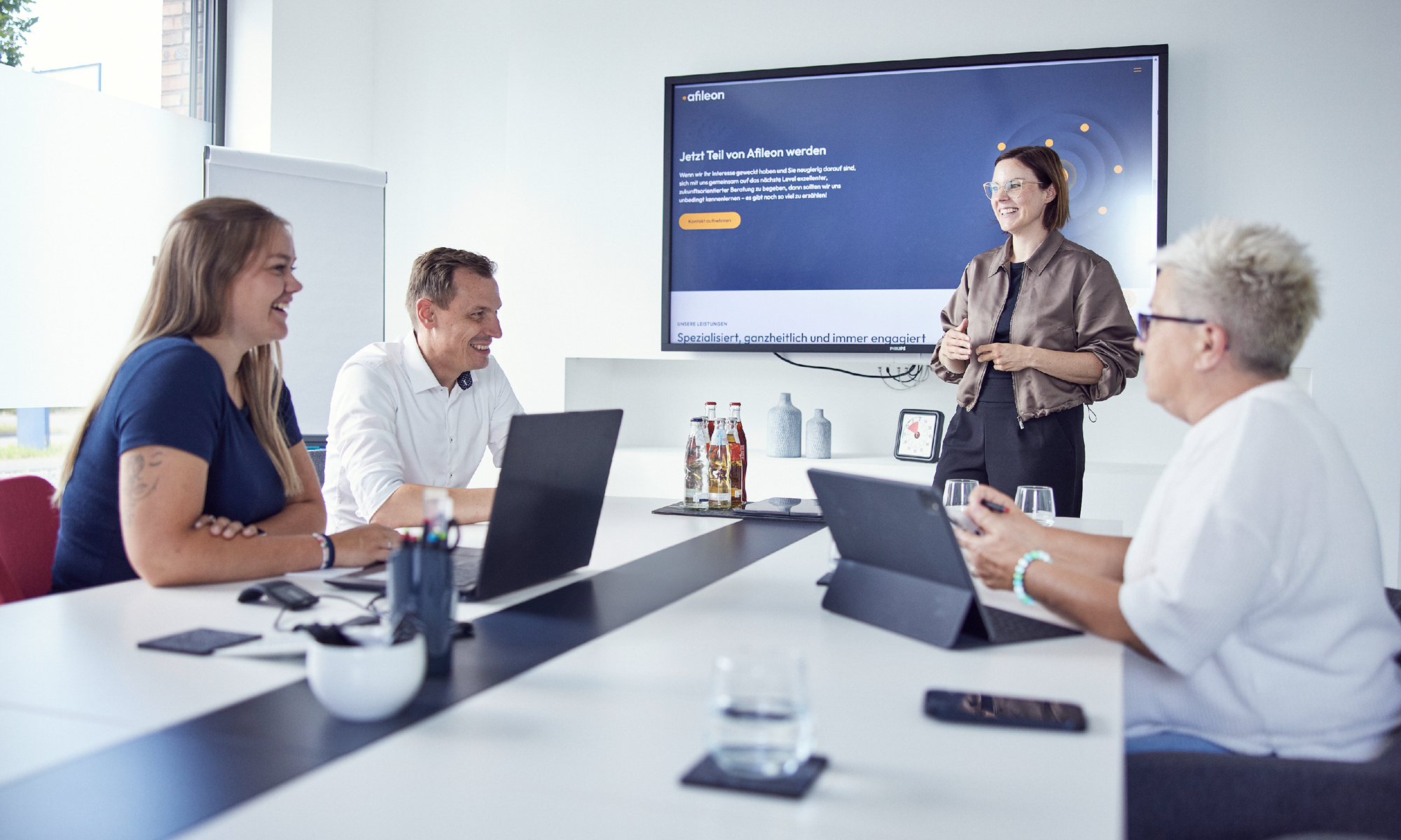 In einem modernen Besprechungsraum präsentieren sich vier Personen während eines Meetings, während eine Frau an der Wand ein digitales Präsentationsmaterial zeigt. Auf dem Tisch stehen Getränke und Laptops.