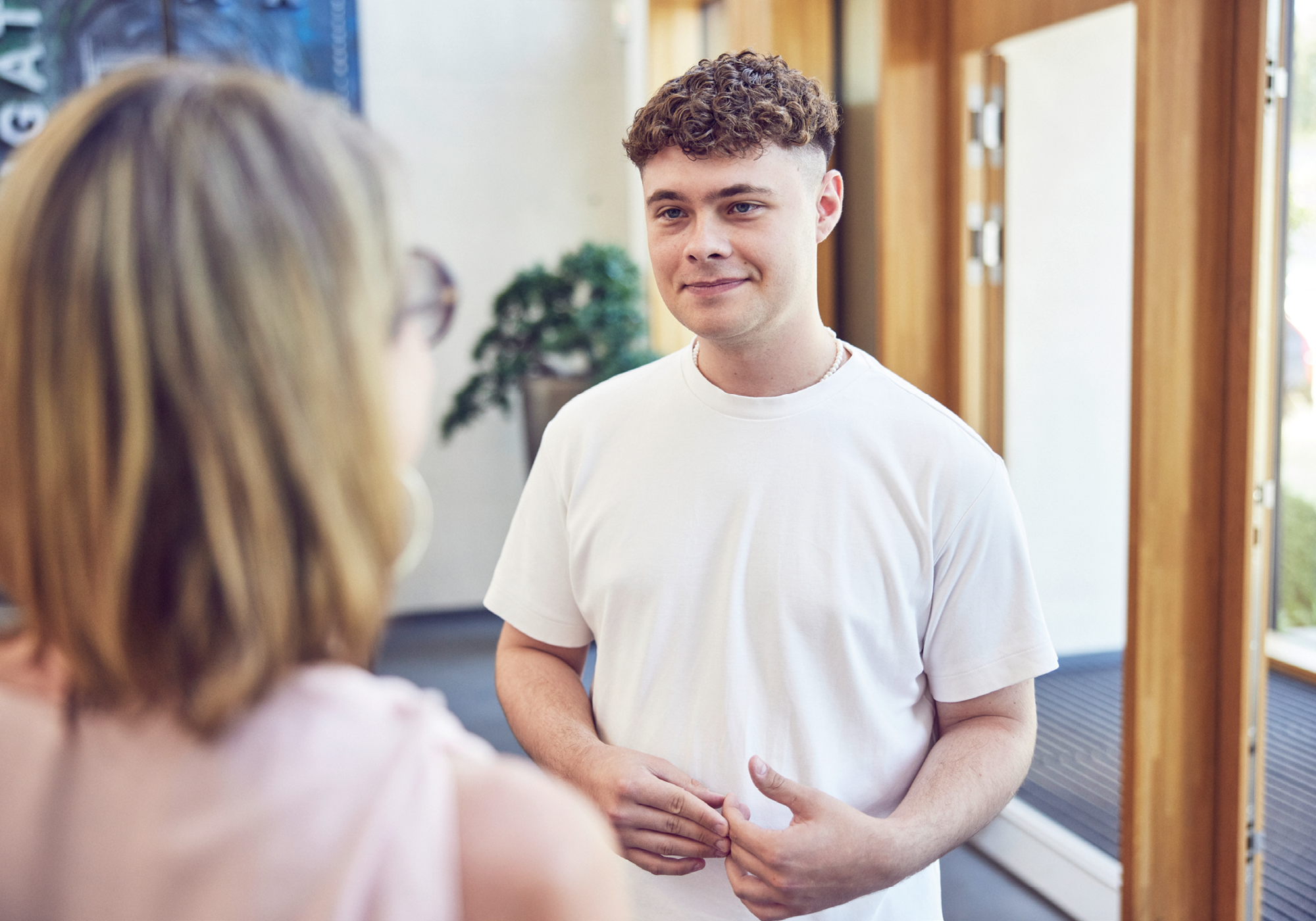 Ein junger Mann mit lockigem, blondem Haar trägt ein einfaches weißes T-Shirt und lächelt, während er mit einer Person spricht, die ihm gegenübersteht. Im Hintergrund sind Holz- und Glasdetails des Raumes zu sehen.