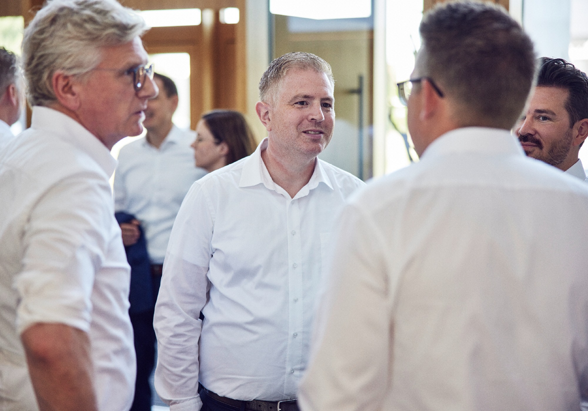 Eine Gruppe von Männern in weißen Hemden unterhält sich in einem hellen, modernen Raum, während im Hintergrund weitere Personen zu sehen sind.