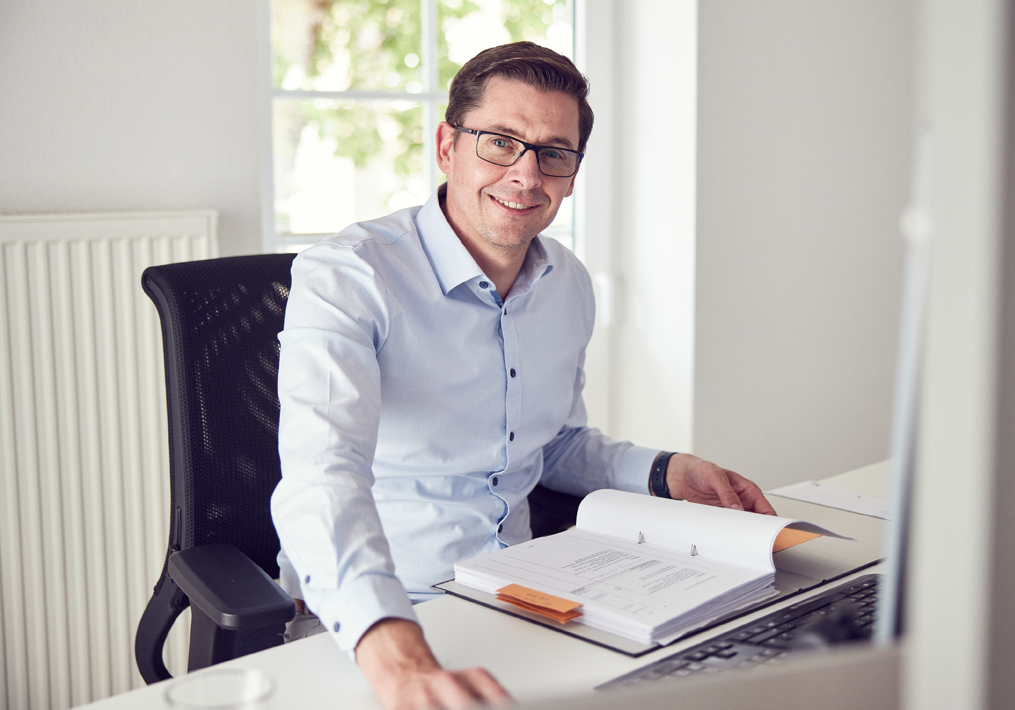 Ein Mann mit Brille sitzt an einem Schreibtisch und lächelt, während er auf ein geöffnetes Dokument blickt. Im Hintergrund sind ein Fenster und eine Heizung zu sehen.