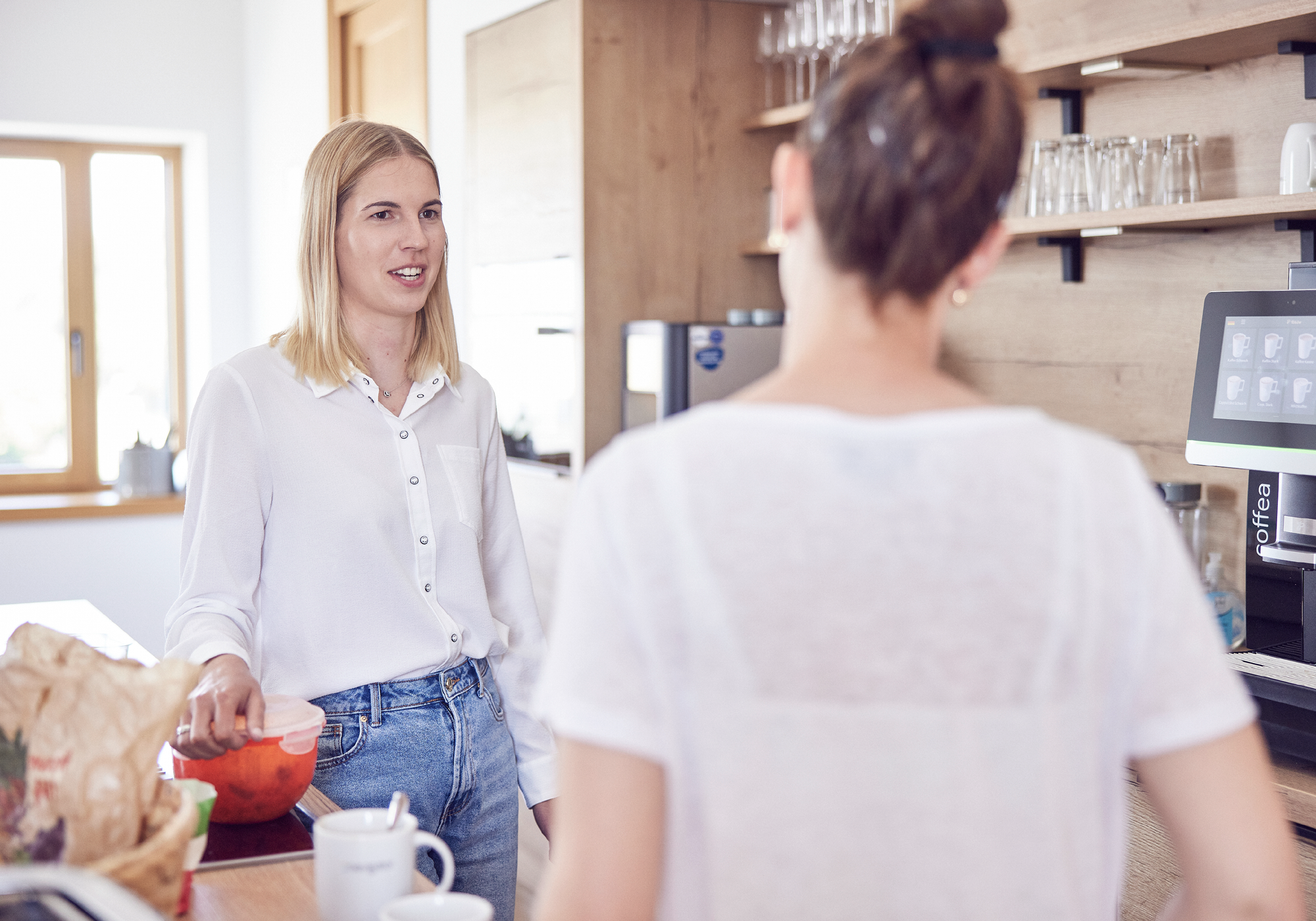 Zwei Frauen stehen in einer modernen Küche; eine Frau spricht mit einer Handbewegung, während die andere Frau ihr zuhört. Auf dem Tisch befinden sich verschiedene Utensilien und ein Glas.