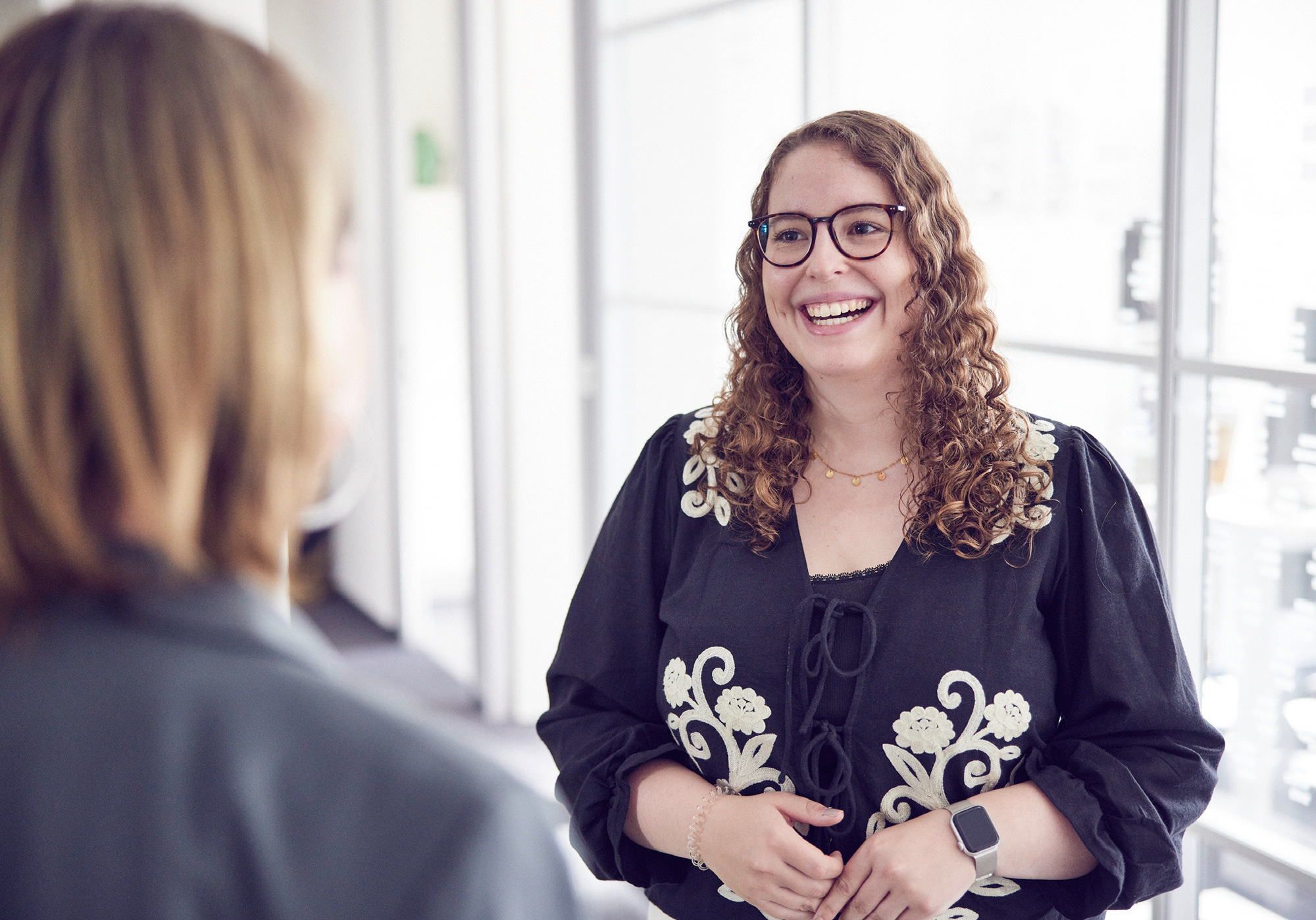 Junge Frau mit Brille lächelt während eines Gesprächs im Büroflur