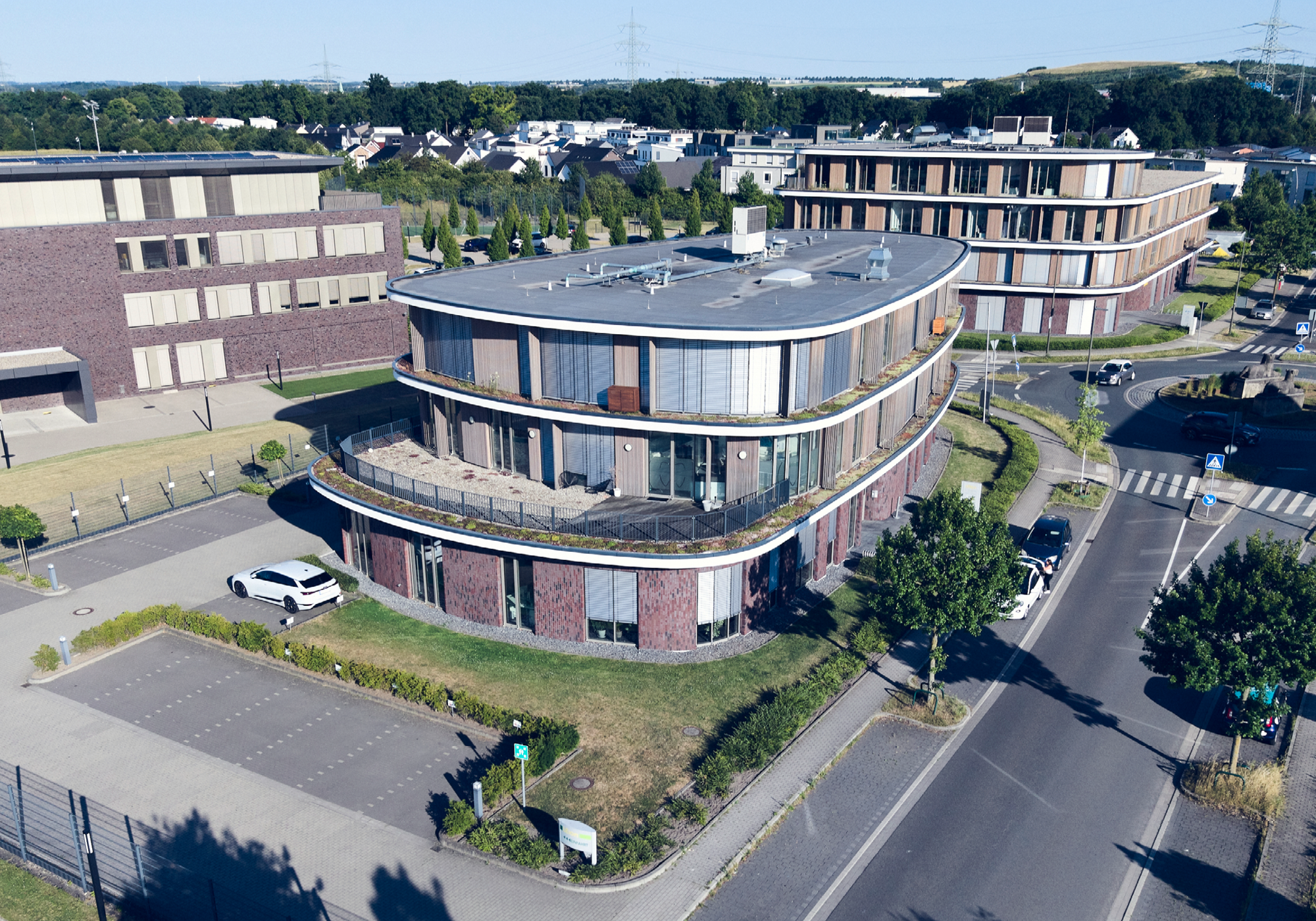 Eine moderne Gebäudestruktur mit geschwungener Fassade und großen Fenstern steht an einer Kreuzung, umgeben von gepflegten Grünflächen und Parkplätzen. Im Hintergrund sind weitere moderne Gebäude und ein Waldgebiet sichtbar.