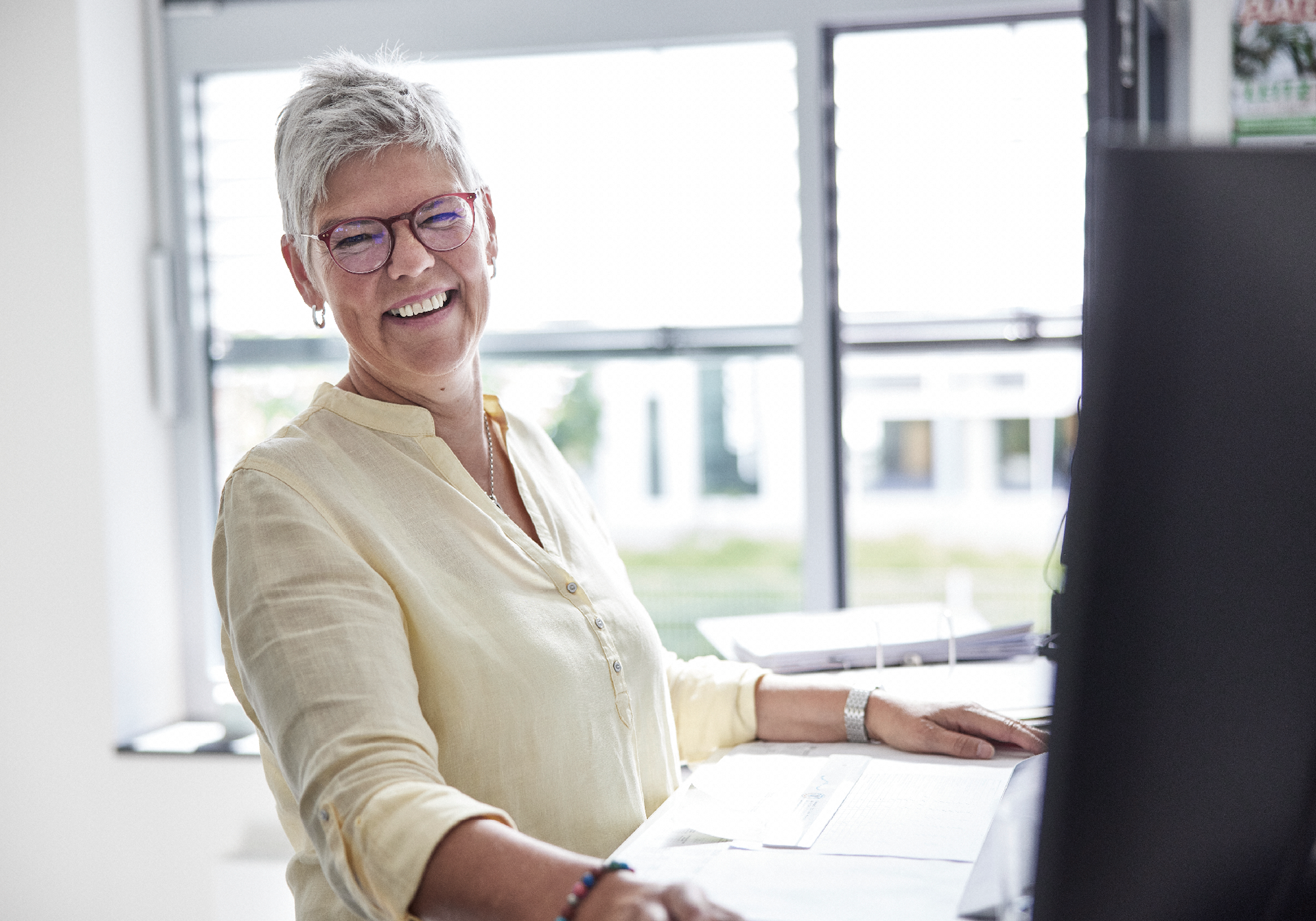 Eine lächelnde Frau mit kurzen grauen Haaren und Brille sitzt an einem Schreibtisch und schaut freundlich in die Kamera. Sie trägt ein hellbeiges Hemd und steht vor einem Computerbildschirm, während im Hintergrund Fenster mit Tageslicht zu sehen sind.