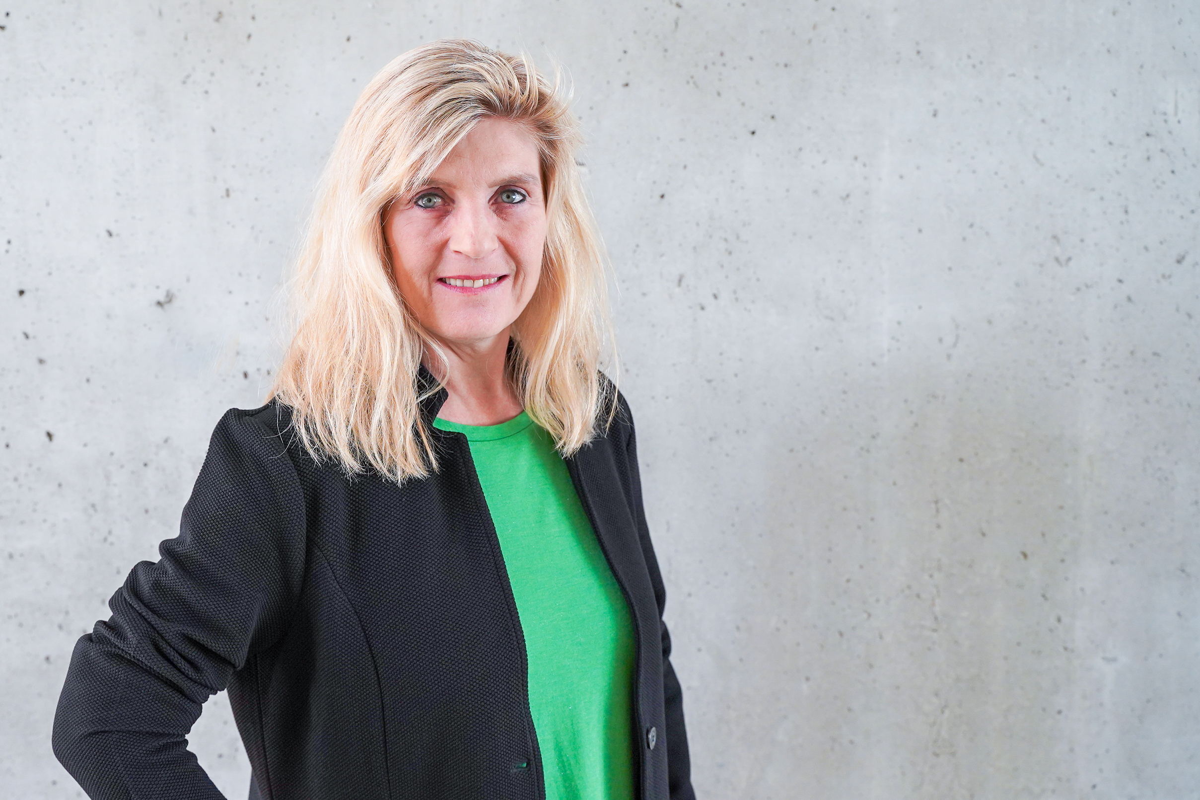 Eine Frau mit langen blonden Haaren trägt ein grünes Shirt und eine schwarze Jacke. Sie steht lächelnd vor einer grauen, strukturierten Wand.