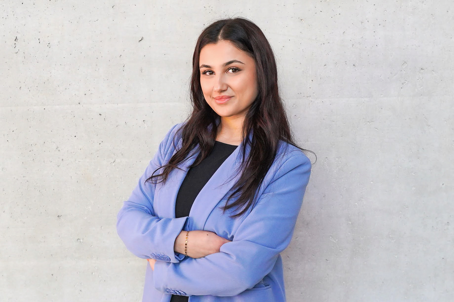 Eine lächelnde Frau in einem lilafarbenen Blazer steht mit verschränkten Armen vor einer grauen Wand.