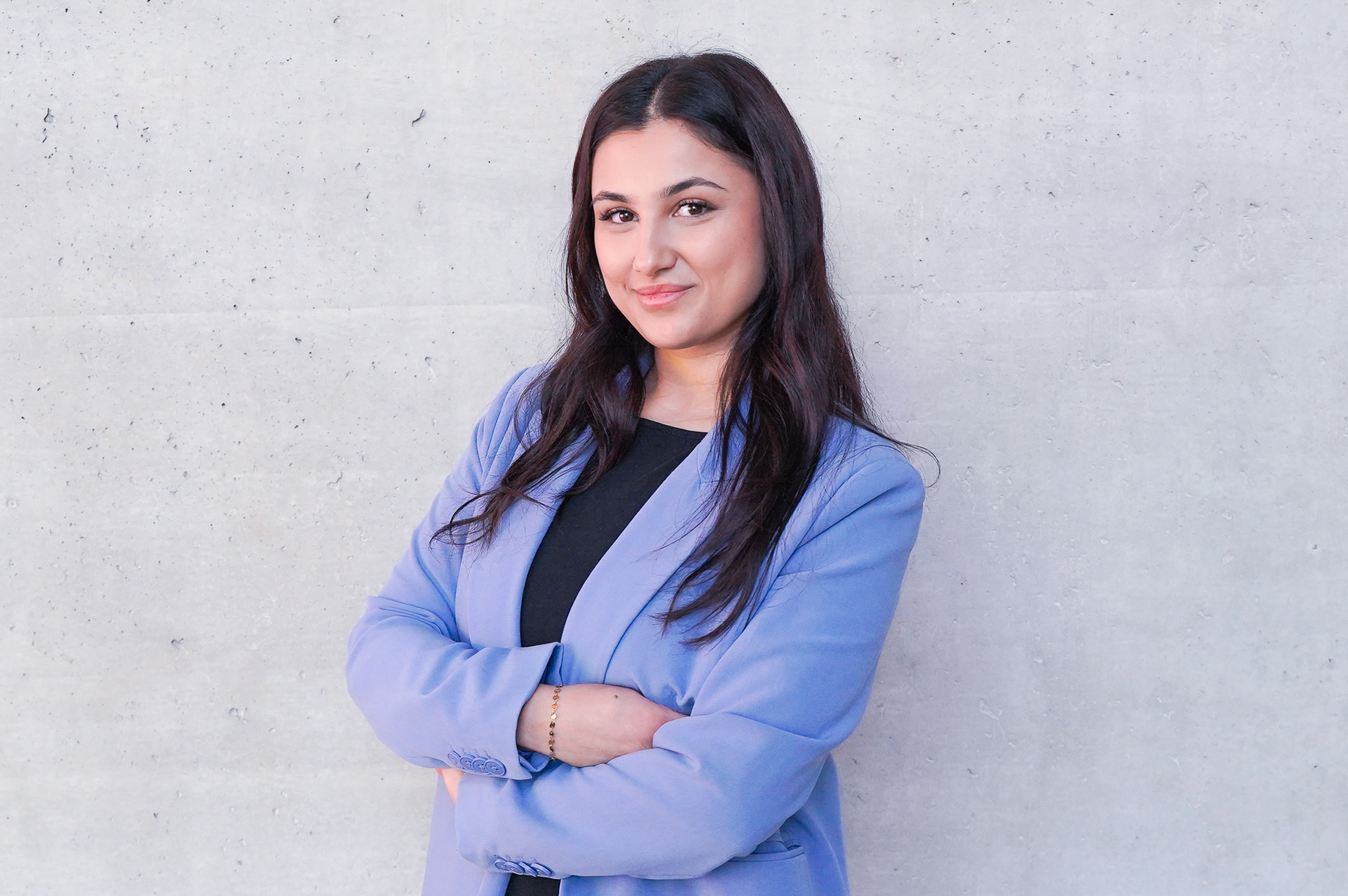 Eine Frau mit langen, dunklen Haaren in einem blauen Blazer steht vor einer grauen Wand und lächelt selbstbewusst, ihre Arme vor der Brust verschränkt.
