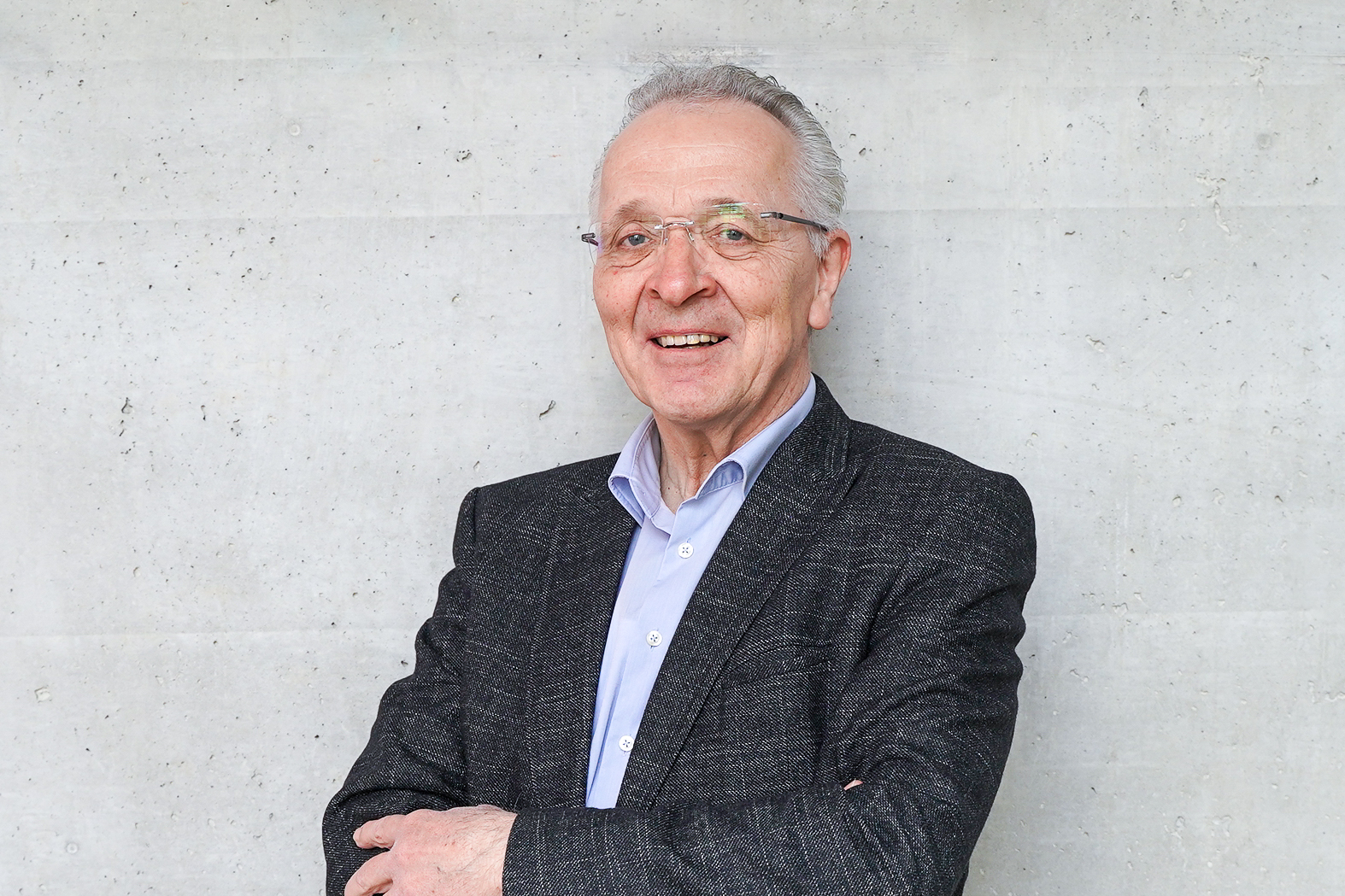 Ein älterer Mann mit grauen Haaren und einer Brille steht lächelnd vor einer grauen Betonwand und trägt einen dunklen Anzug.
