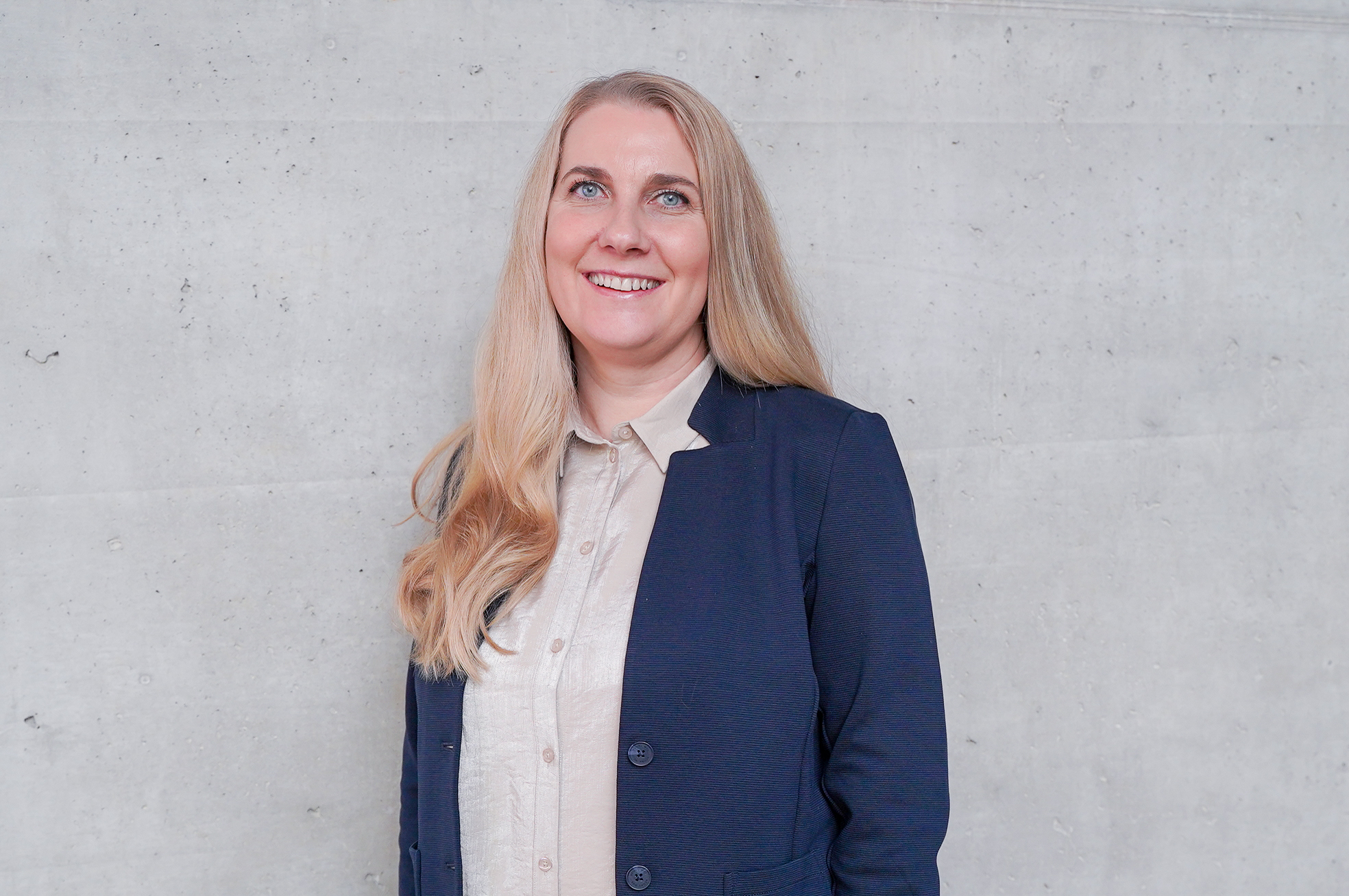 Eine blonde Frau mit langen, glatten Haaren lächelt vor einer ungefähren grauen Betonwand. Sie trägt ein helles Hemd und einen dunklen Blazer.