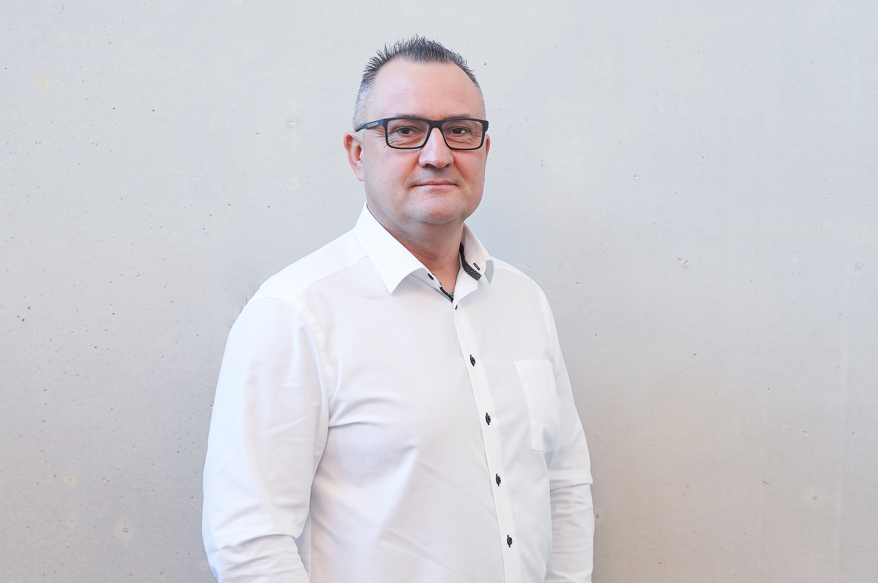 Ein Mann mit Brille und kurzen, grauen Haaren trägt ein weißes Hemd mit schwarzen Knöpfen und steht vor einem hellen, einfarbigen Hintergrund.