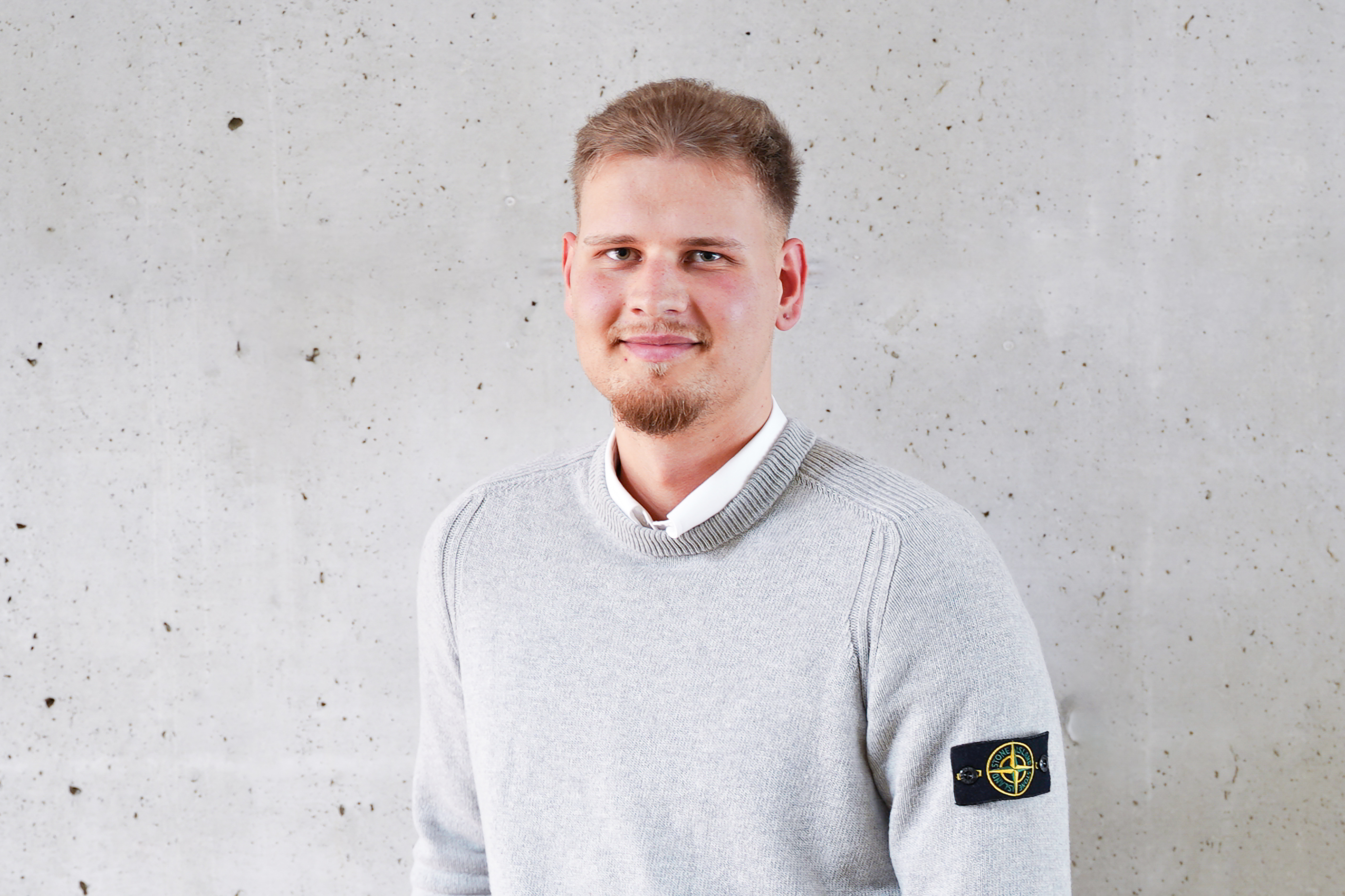 Ein junger Mann mit kurzen, blonden Haaren und einem leichten Bart trägt einen grau gestrickten Pullover mit einem Patch auf dem linken Ärmel. Er steht vor einer grauen Wand mit einer strukturierten Oberfläche und lächelt freundlich.