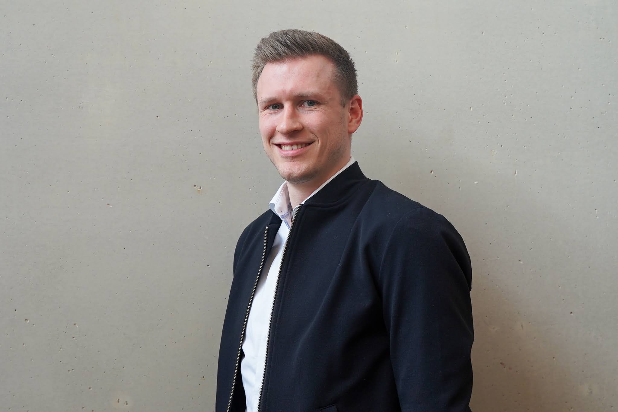 Eine Person mit kurzen Haaren und einem freundlichen Lächeln steht vor einer grauen Wand und trägt eine dunkle Jacke über einem weißen Hemd.