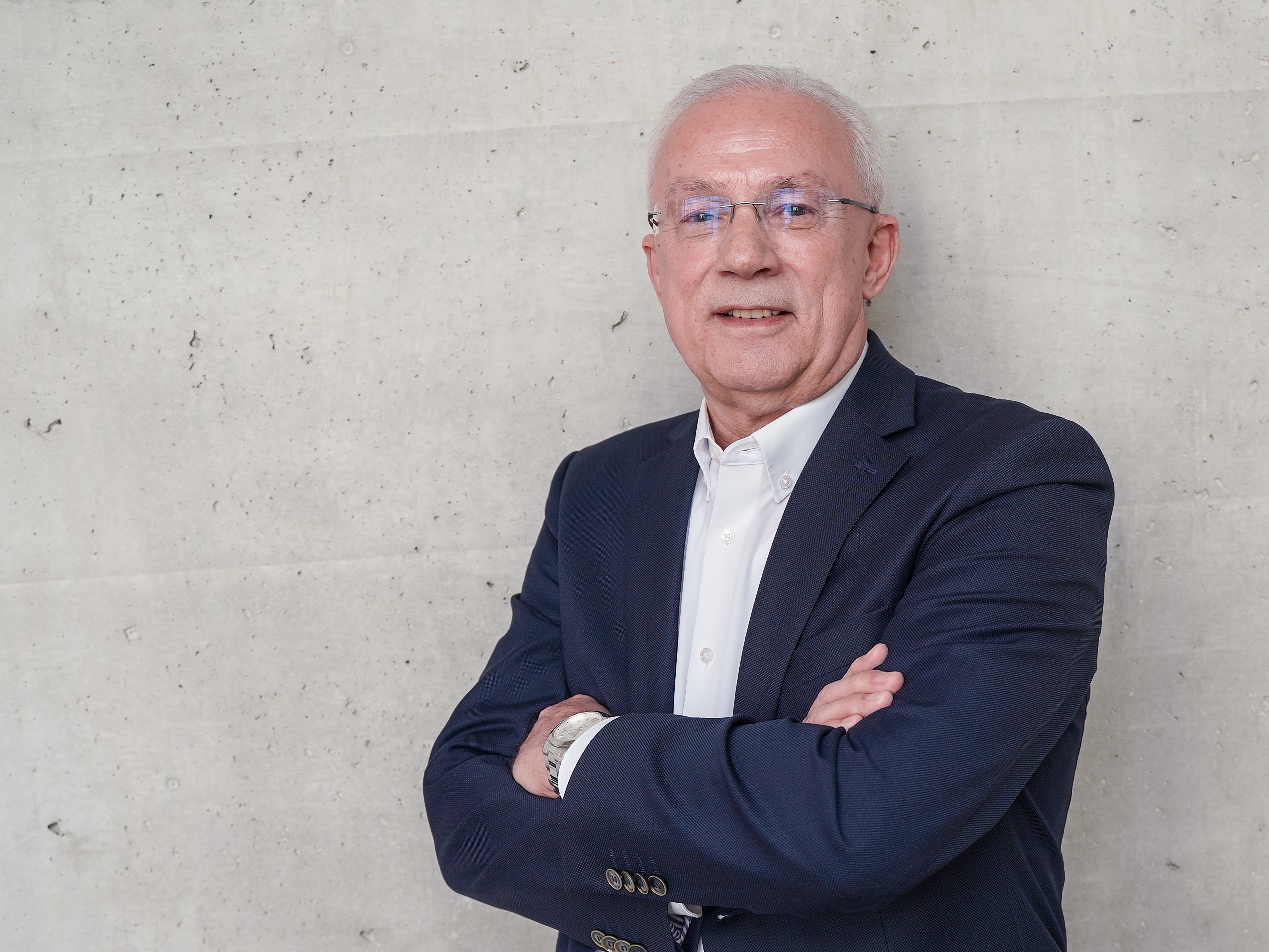 Ein älterer Mann mit grauen Haaren und Brille steht lächelnd mit verschränkten Armen vor einer grauen, strukturierten Wand. Er trägt ein dunkelblaues Sakko und ein weißes Hemd.