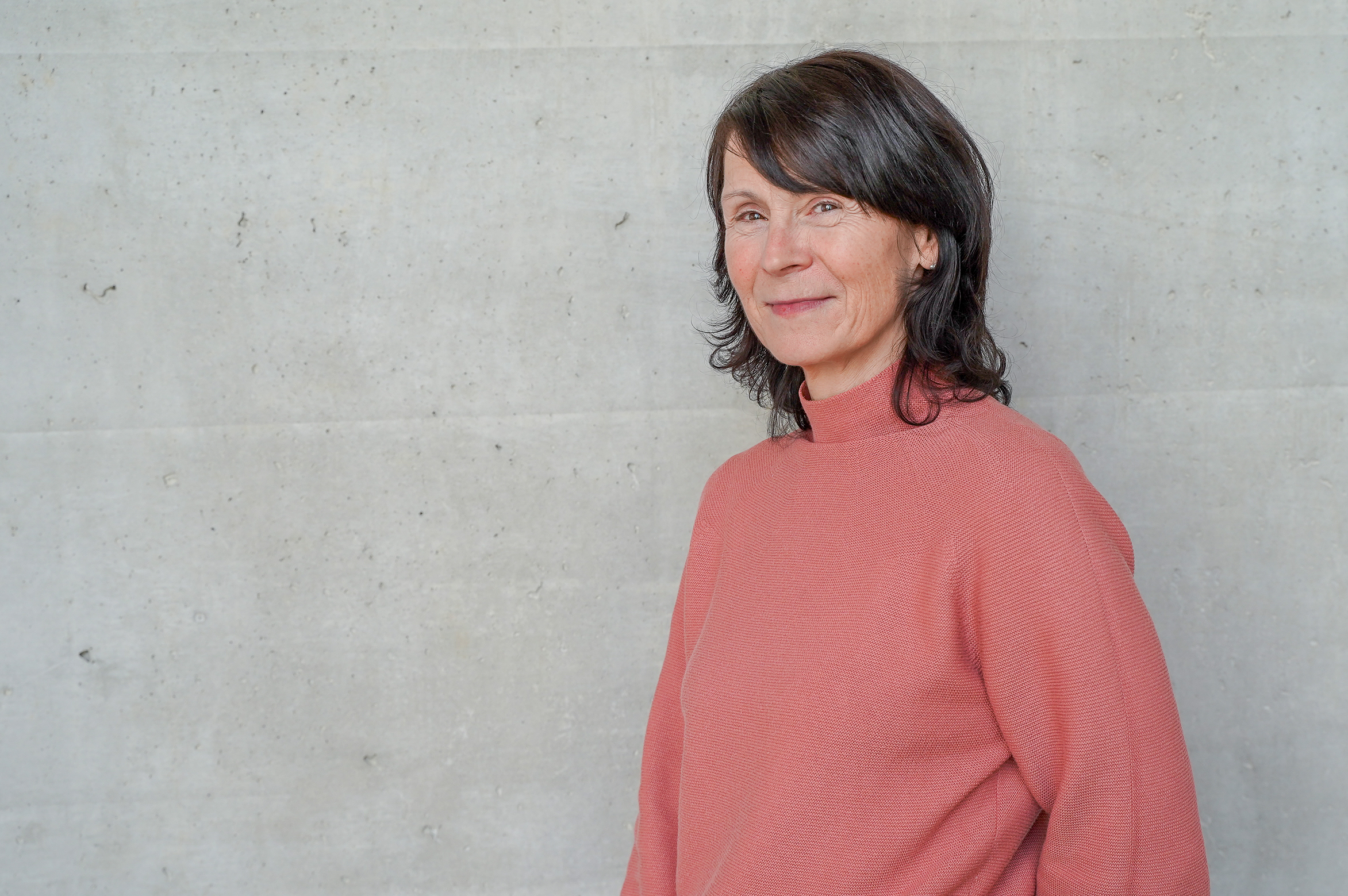 Eine lächelnde Frau mit langen, dunklen Haaren steht seitlich vor einer grauen, strukturierten Wand und trägt einen rosa Pullover.