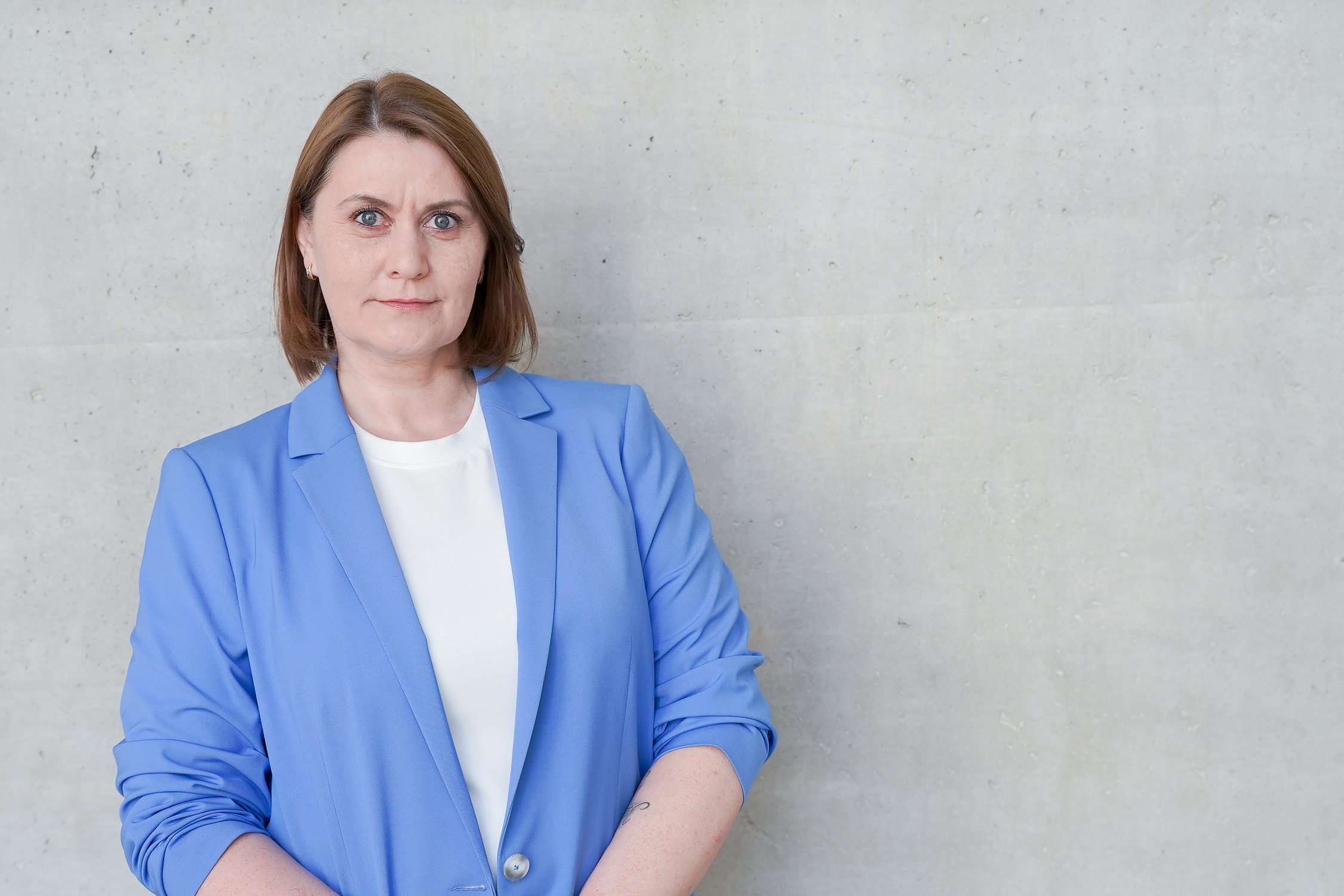 Eine Person mit kurzen braunen Haaren trägt einen blauen Blazer und schaut direkt in die Kamera, vor einer hellgrauen Wand.
