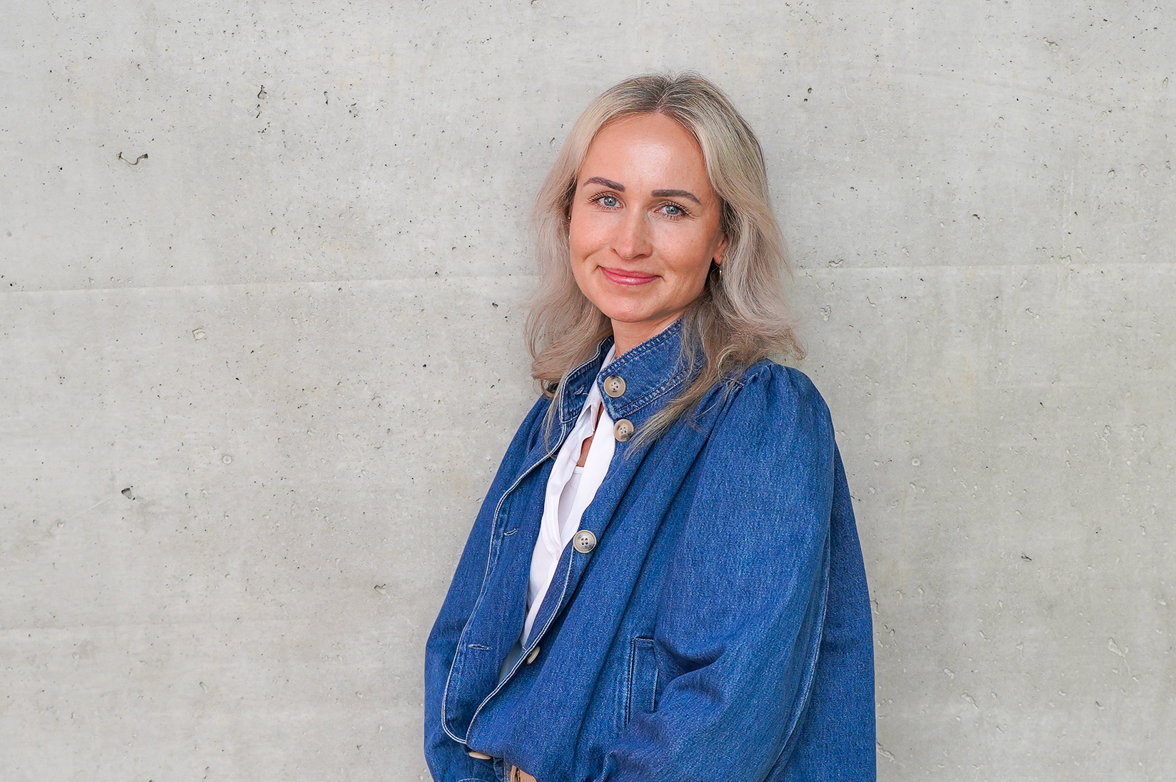 Eine Frau mit blonden Haaren trägt eine blaue Denim-Jacke und steht vor einer neutralen, grauen Wand. Sie lächelt freundlich in die Kamera.