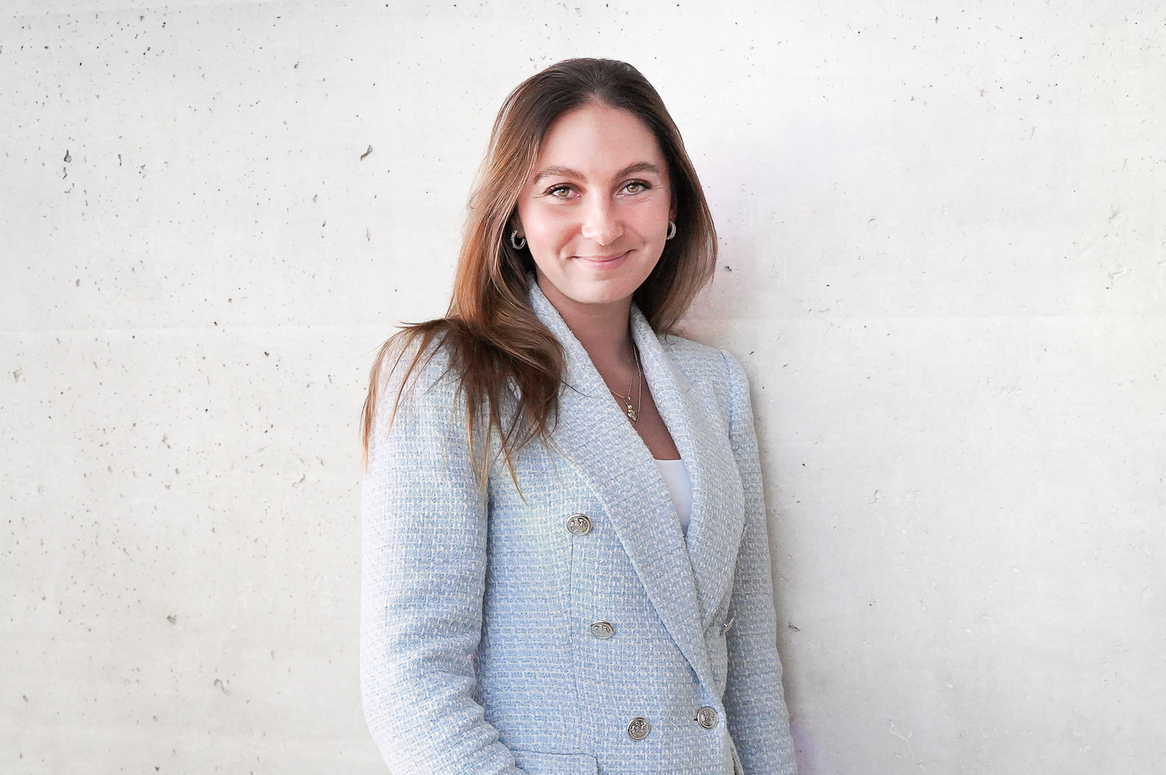 Eine lächelnde Frau mit langen, braunen Haaren trägt einen hellen, zweireihigen Blazer und steht neben einer hellen, strukturierten Wand.