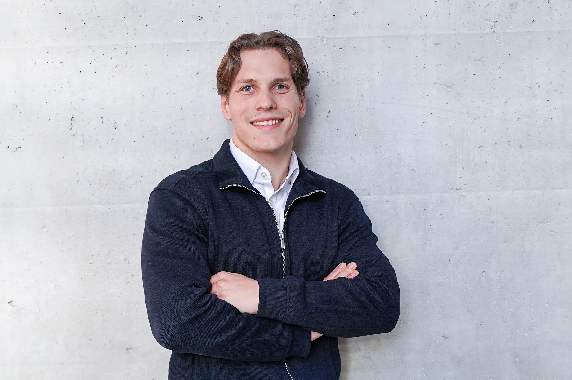 Ein junger Mann mit kurzen, lockigen Haaren steht lächelnd vor einer grauen Betonwand, während er die Arme vor der Brust gekreuzt hat und einen schwarzen Pullover trägt.