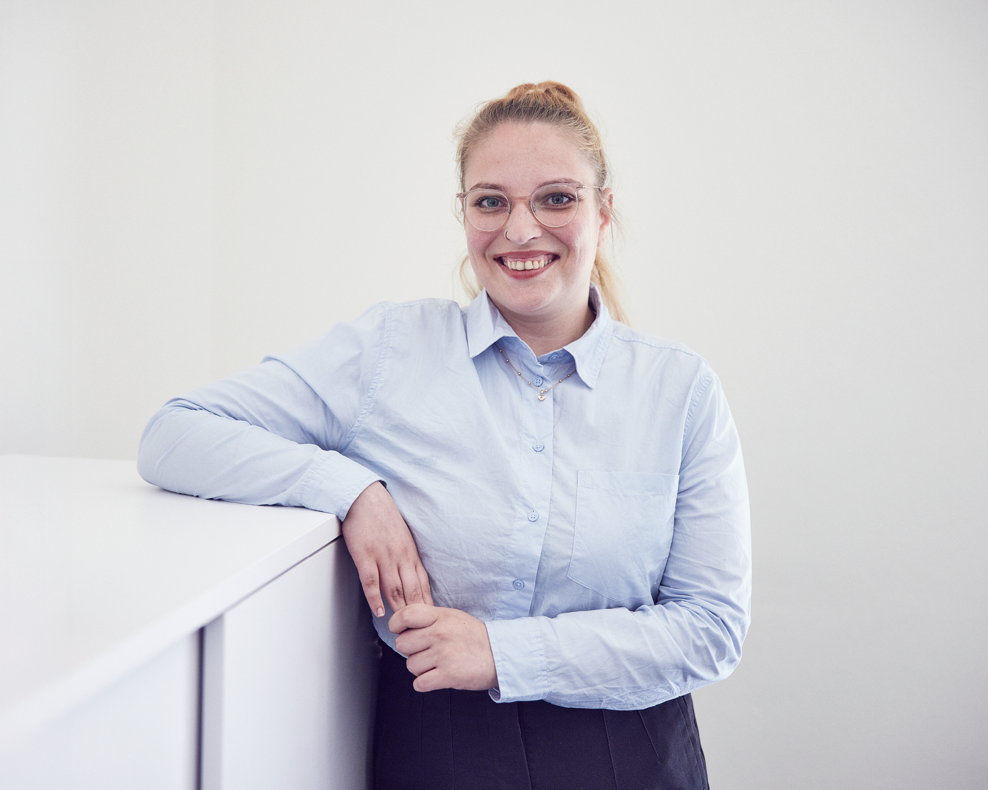 Eine lächelnde Frau mit blonden Haaren, die zu einem Pferdeschwanz gebunden sind, trägt eine hellblaue Bluse und steht entspannt an einem weißen Regal in einem hellen Raum.