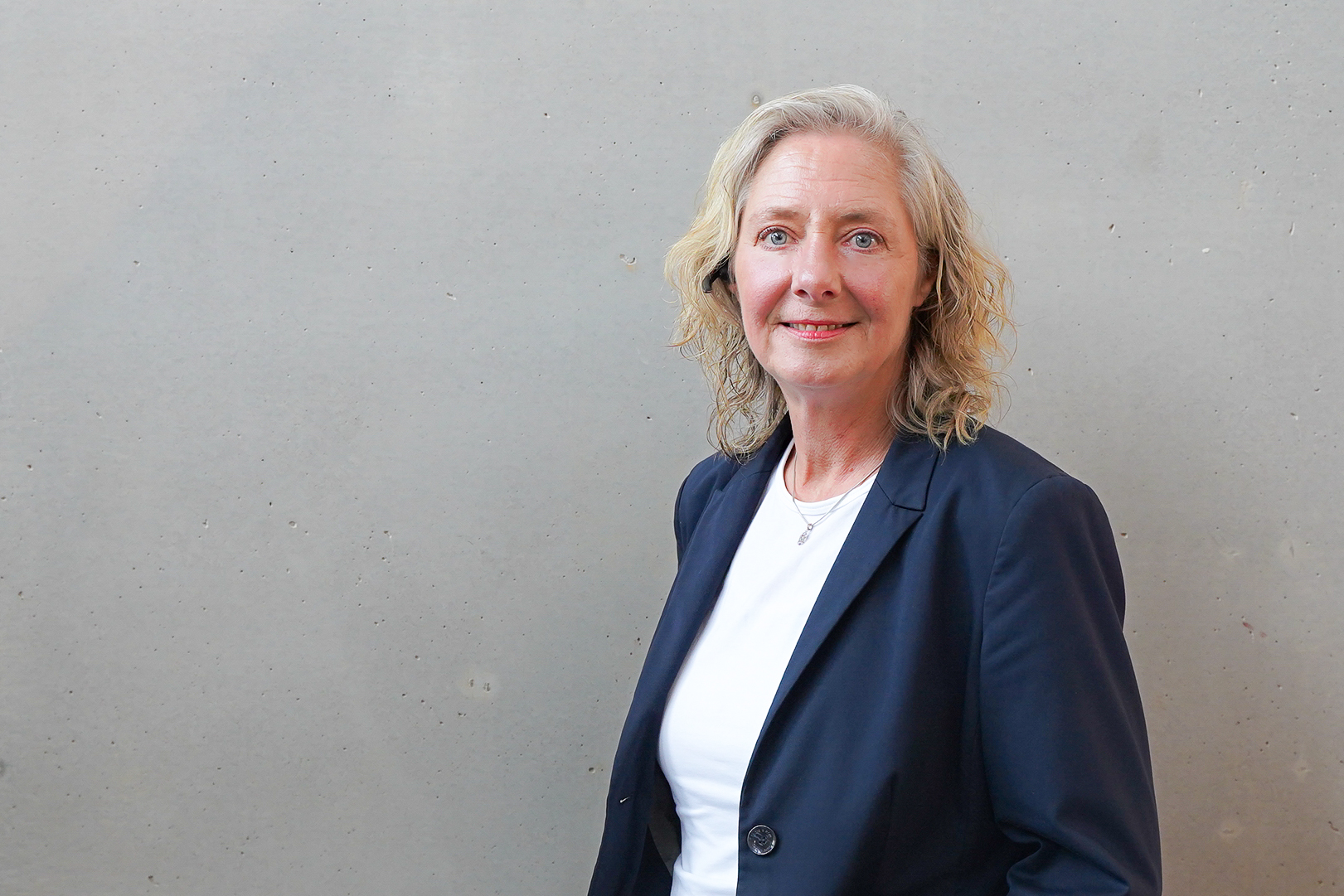 Eine lächelnde Frau mit lockigem, blondem Haar steht vor einer grauen Wand und trägt einen dunkelblauen Blazer über einem weißen Oberteil.