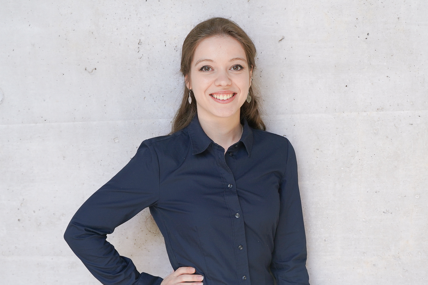 Eine lächelnde junge Frau steht mit einer Hand in die Hüfte gestützt vor einer grauen Wand. Sie trägt ein dunkles Hemd und hat ihre Haare offen.