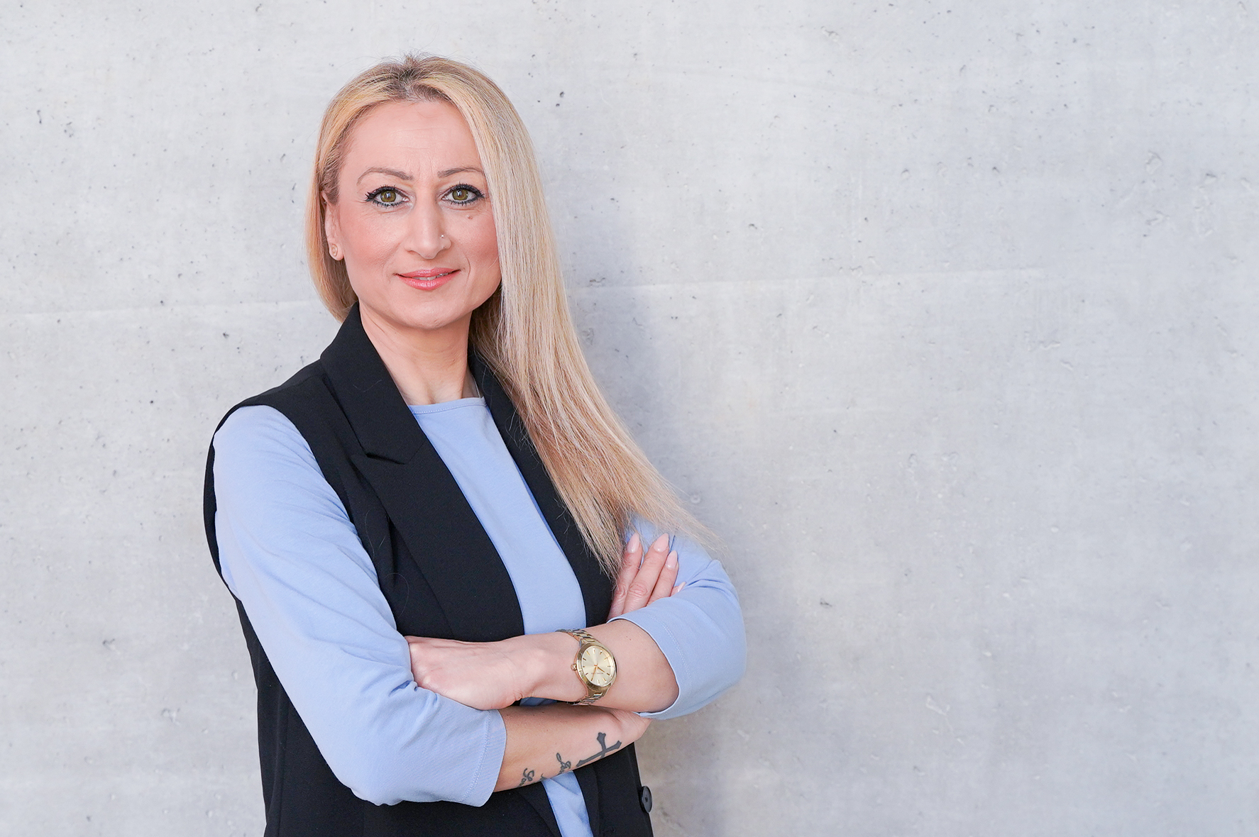 Eine Person mit langen blonden Haaren steht mit überkreuzten Armen vor einer grauen, strukturierten Wand. Sie trägt ein hellblaues Oberteil und einen schwarzen Blazer, wobei sie freundlich in die Kamera lächelt.