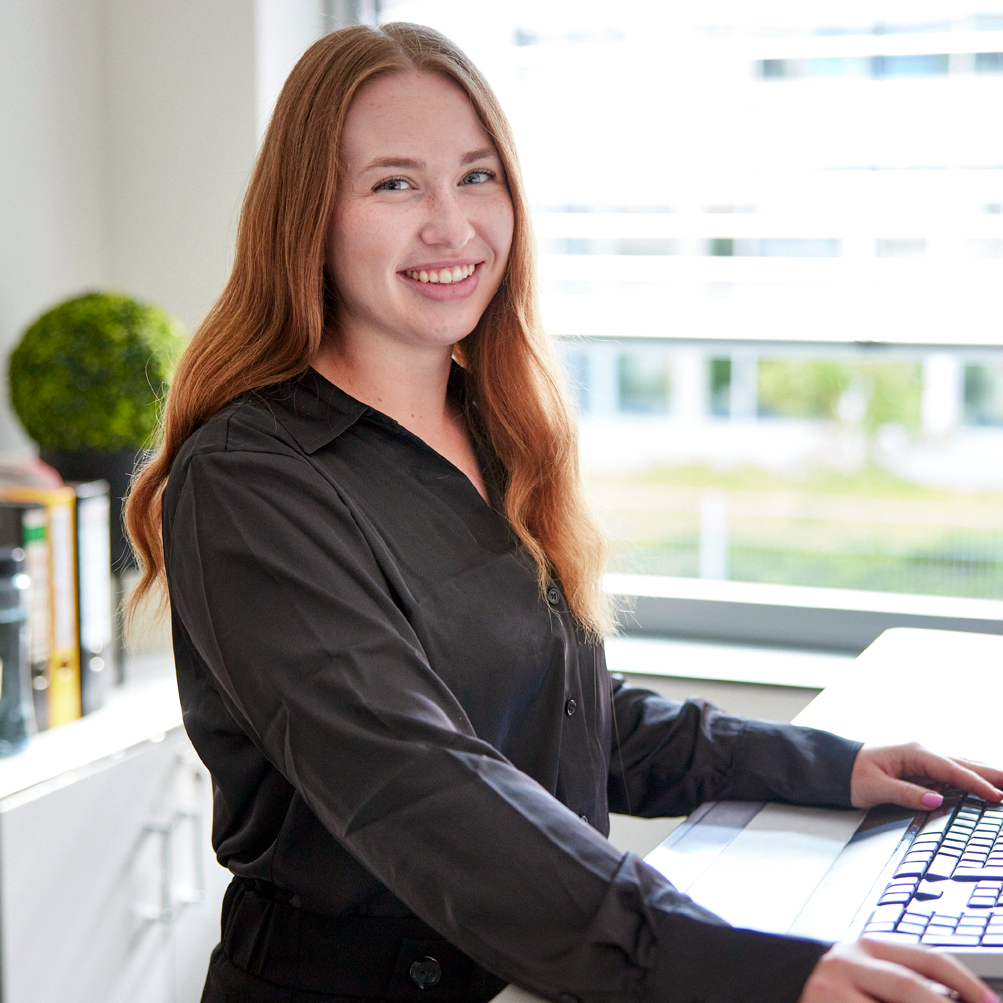 Eine lächelnde Frau mit langen, lockigen roten Haaren steht an einem Arbeitsplatz und tippt auf einer Computertastatur, während sie in die Kamera schaut. Im Hintergrund ist ein Fenster mit natürlichem Licht und eine grüne Pflanze sichtbar.