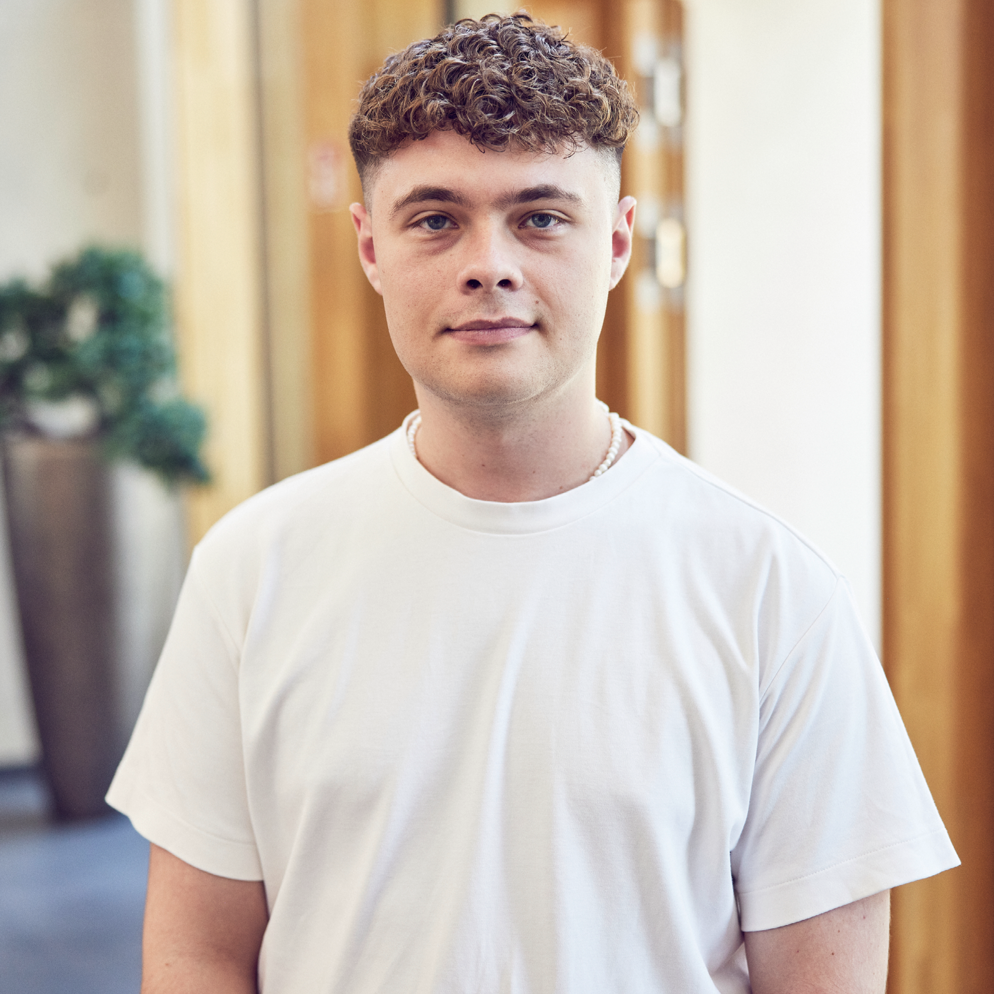 Ein junger Mann mit lockigem, blondem Haar trägt ein einfaches weißes T-Shirt und sieht direkt in die Kamera. Im Hintergrund sind unscharf eine Pflanze und Holzpaneele zu erkennen.