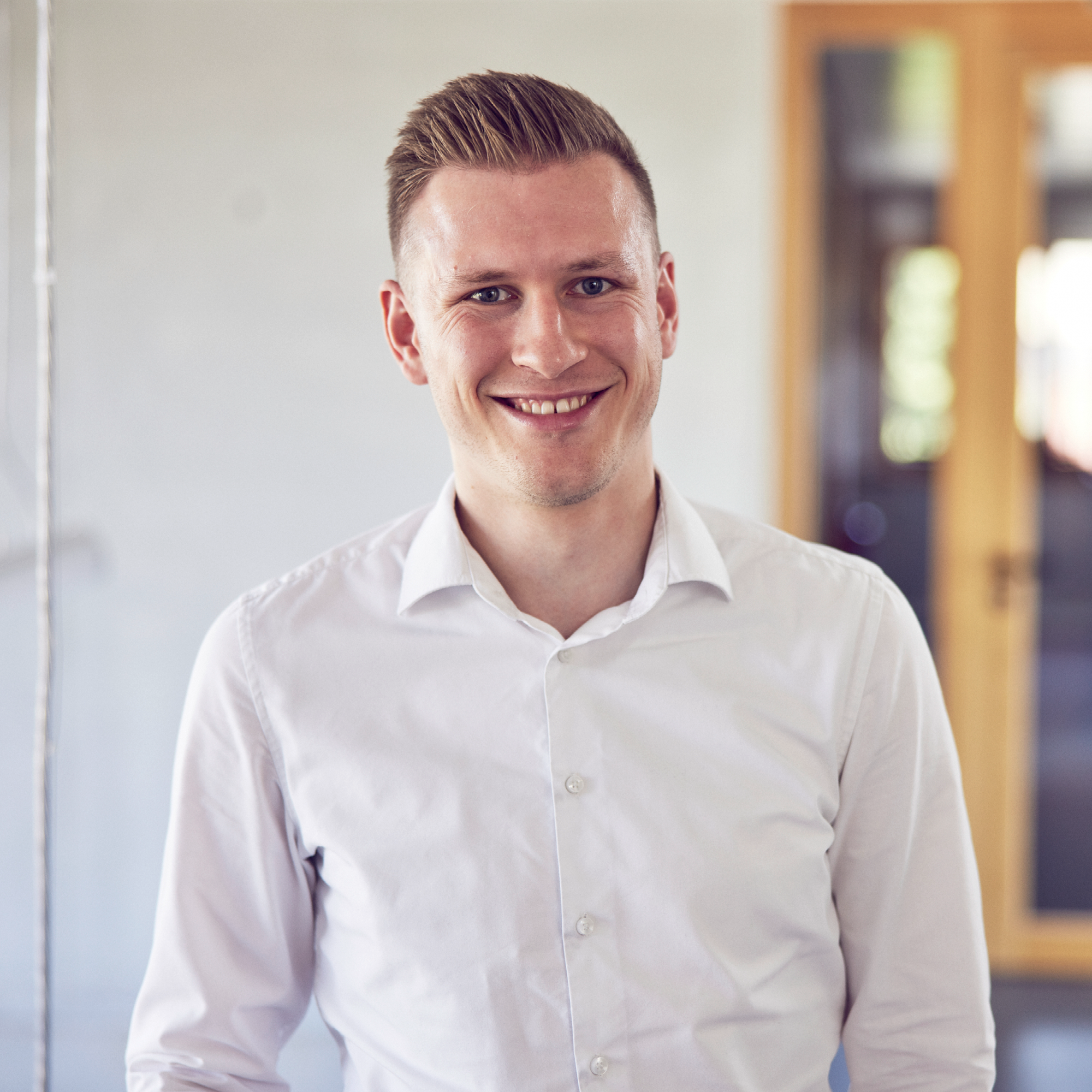 Ein lächelnder Mann steht in einem modernen, hellen Raum, trägt ein weißes Hemd und hat kurze, gepflegte Haare. Im Hintergrund sind verschwommen Fenster und eine Tür zu sehen.