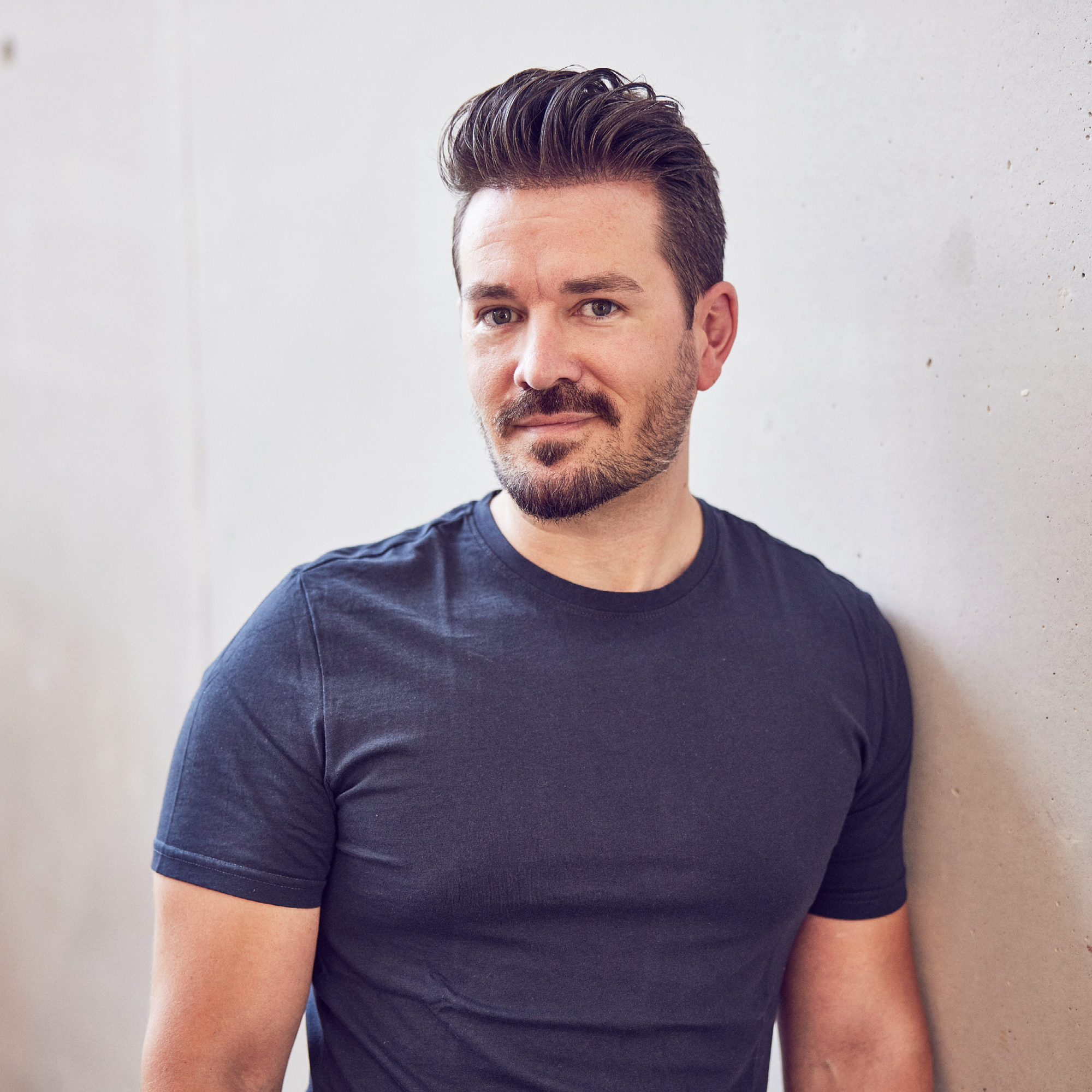 Ein Mann mit kurzen, gestylten Haaren und einem Drei-Tage-Bart steht an einer glatten, hellen Wand und schaut direkt in die Kamera. Er trägt ein schlichtes, dunkelblaues T-Shirt.