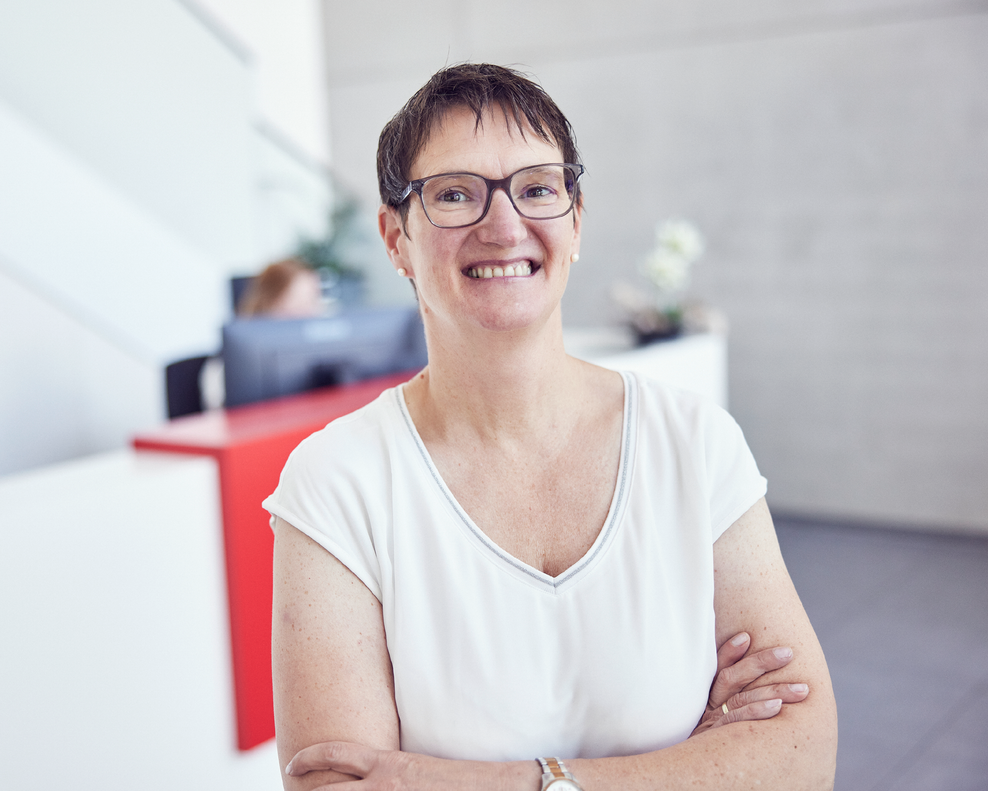 Eine lächelnde Frau mit kurzen, braunen Haaren und Brille steht mit verschränkten Armen in einem modernen Empfangsbereich, während sie in die Kamera schaut. Im Hintergrund ist ein Empfangstresen sichtbar.